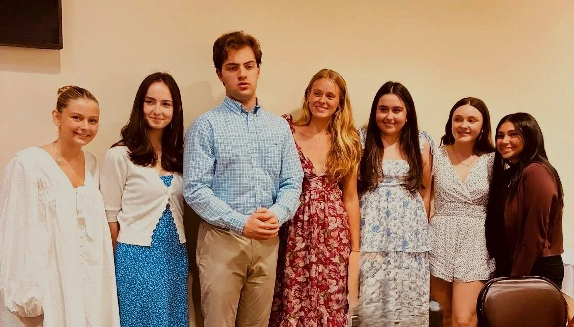 Group of seven young people, six women and one man, standing together indoors, dressed in casual to semi-formal attire, smiling and posing for a photo.