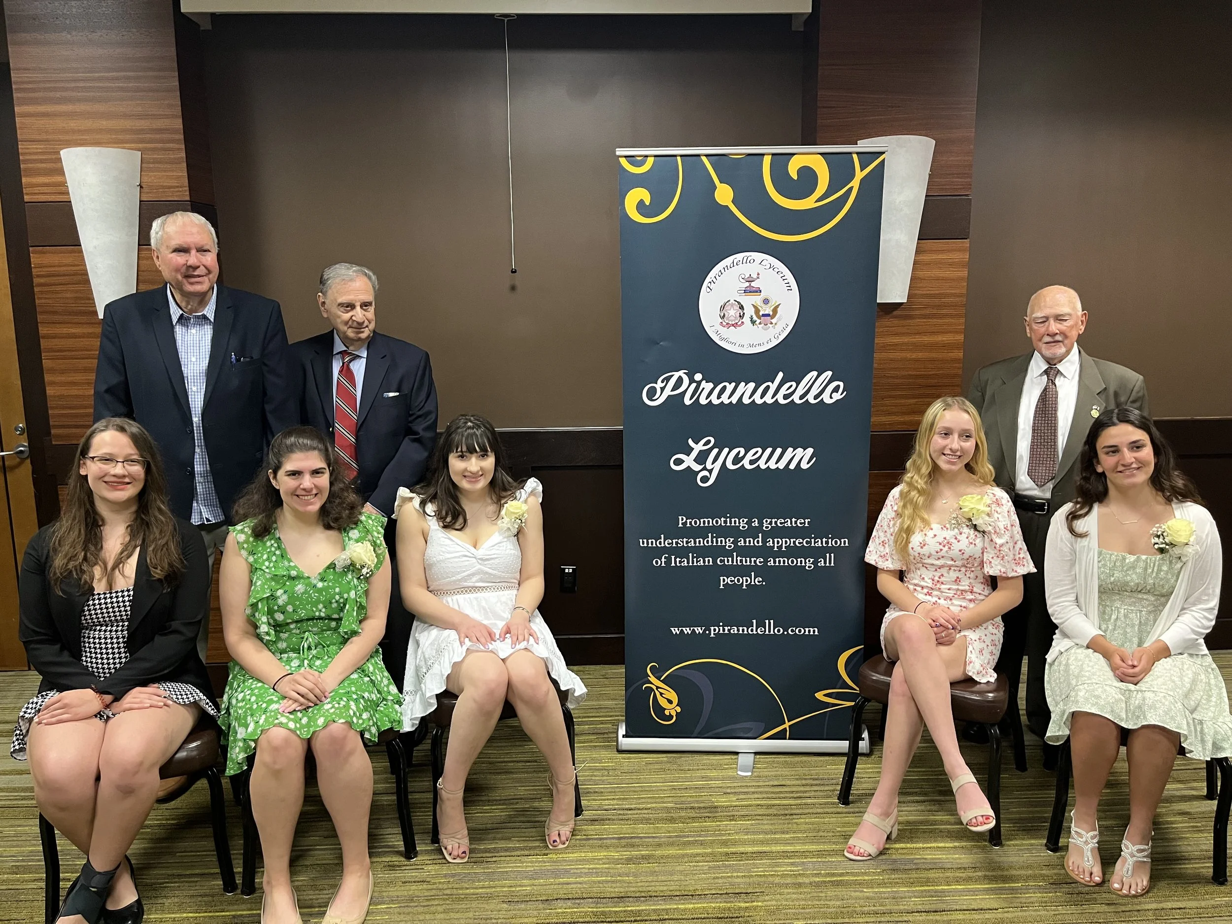 Group photo of six young women and four older men at Pirandello Lyceum event. Women are seated in front, wearing dresses, while men stand behind them, dressed in suits. A blue banner with the Pirandello Lyceum logo and mission statement stands between the groups. The setting appears to be a conference room or banquet hall with wood-paneled walls and carpeted floor.
