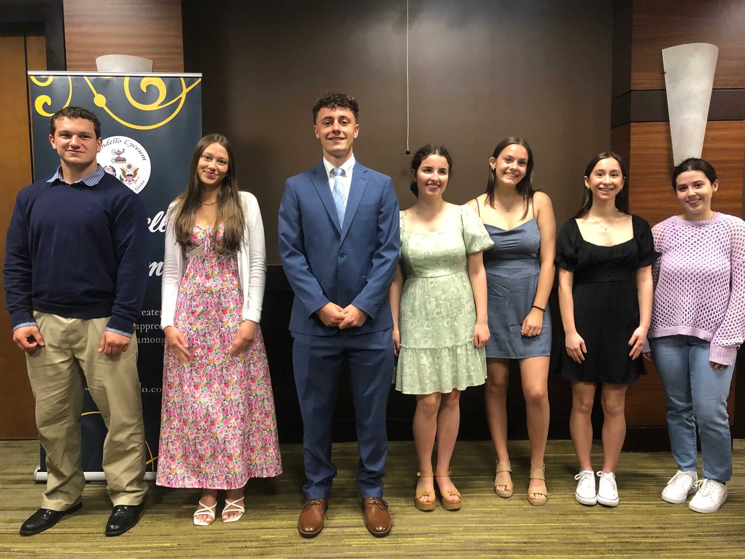 Group of eight young people standing in a line indoors, dressed in semi-formal to casual attire, smiling at the camera, with a banner in the background.
