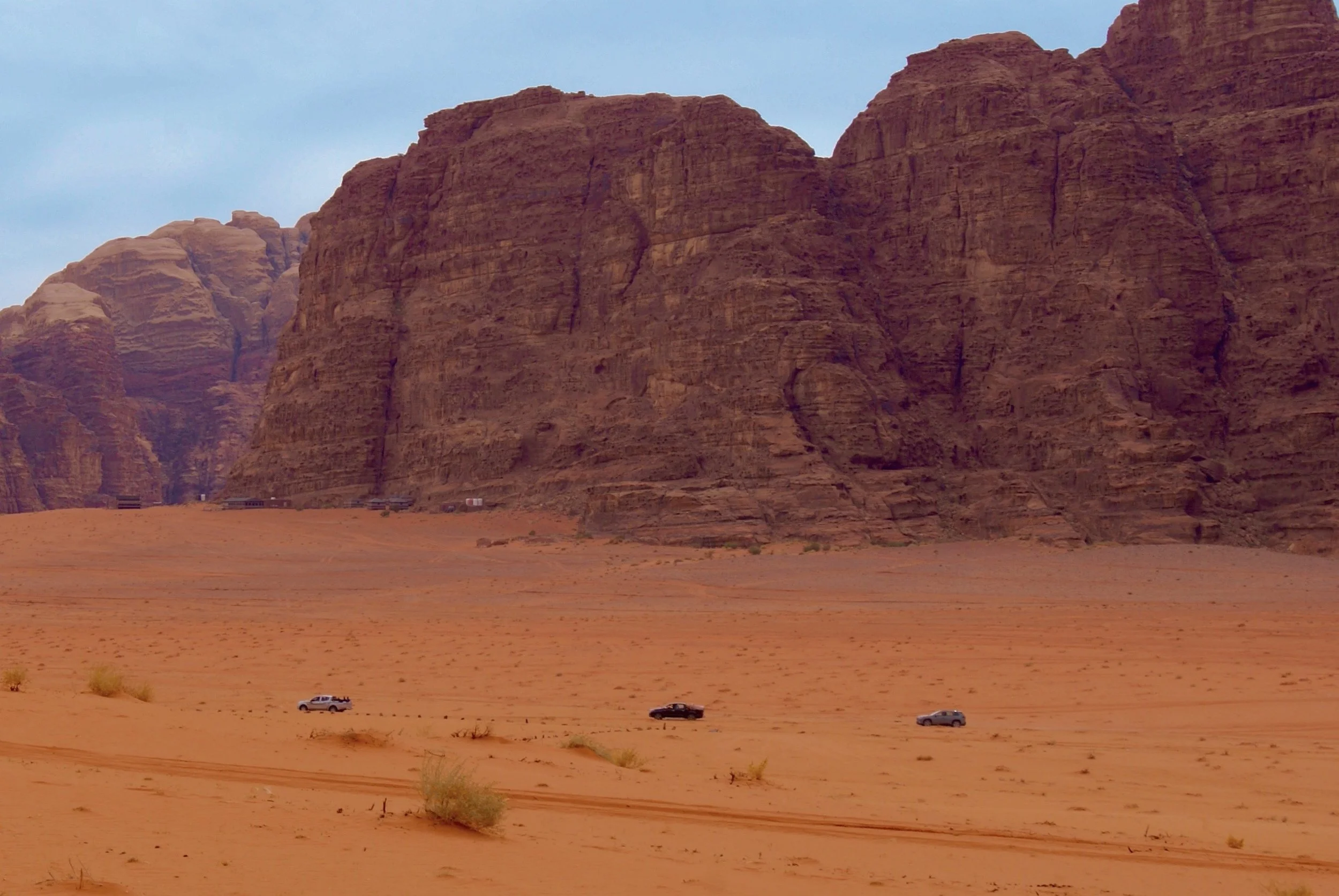 Red desert landscape with large rock formations and a few cars driving on the sandy terrain.