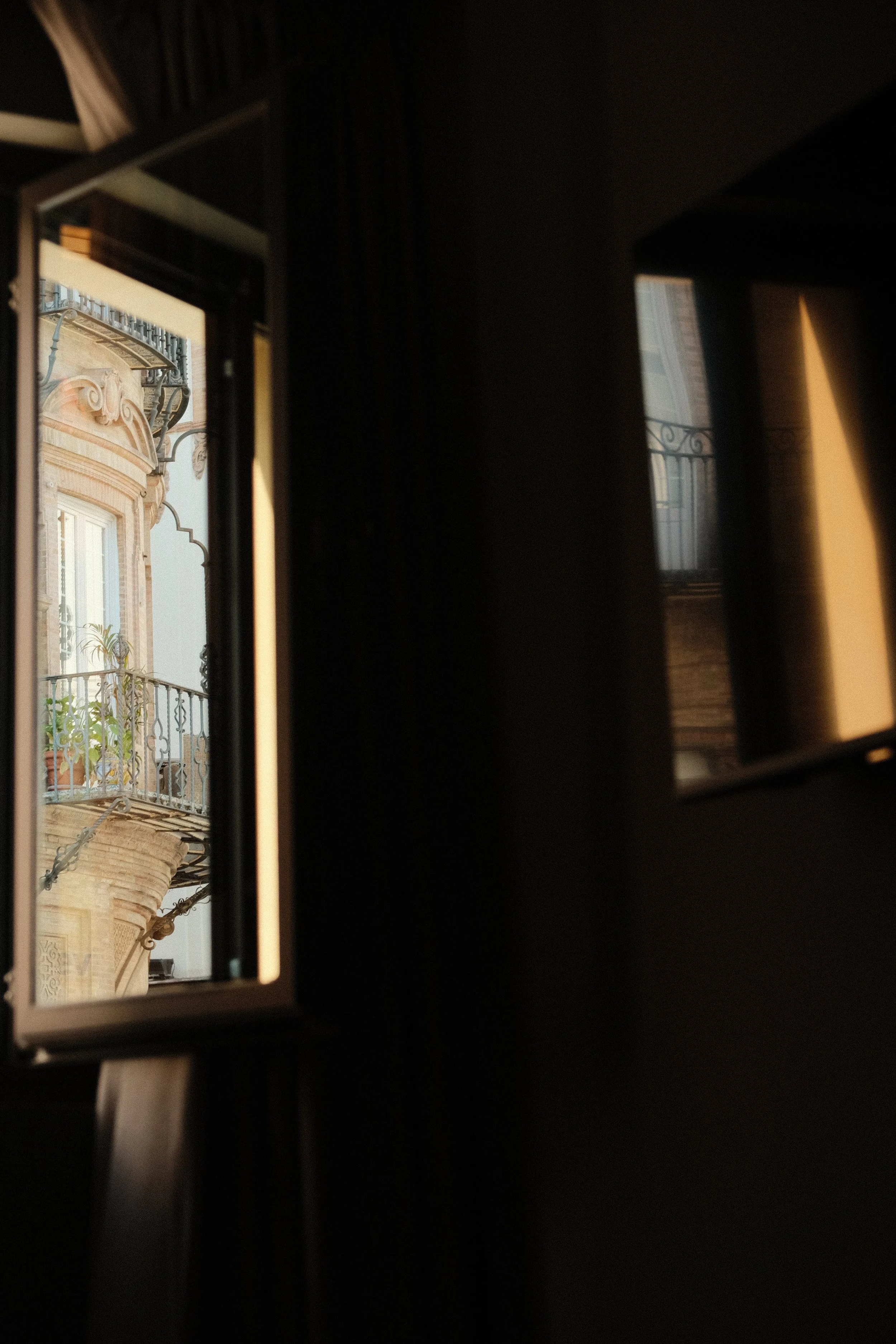 The photo shows a view through two small open windows, revealing an ornate building with decorative iron balconies and potted plants outside.
