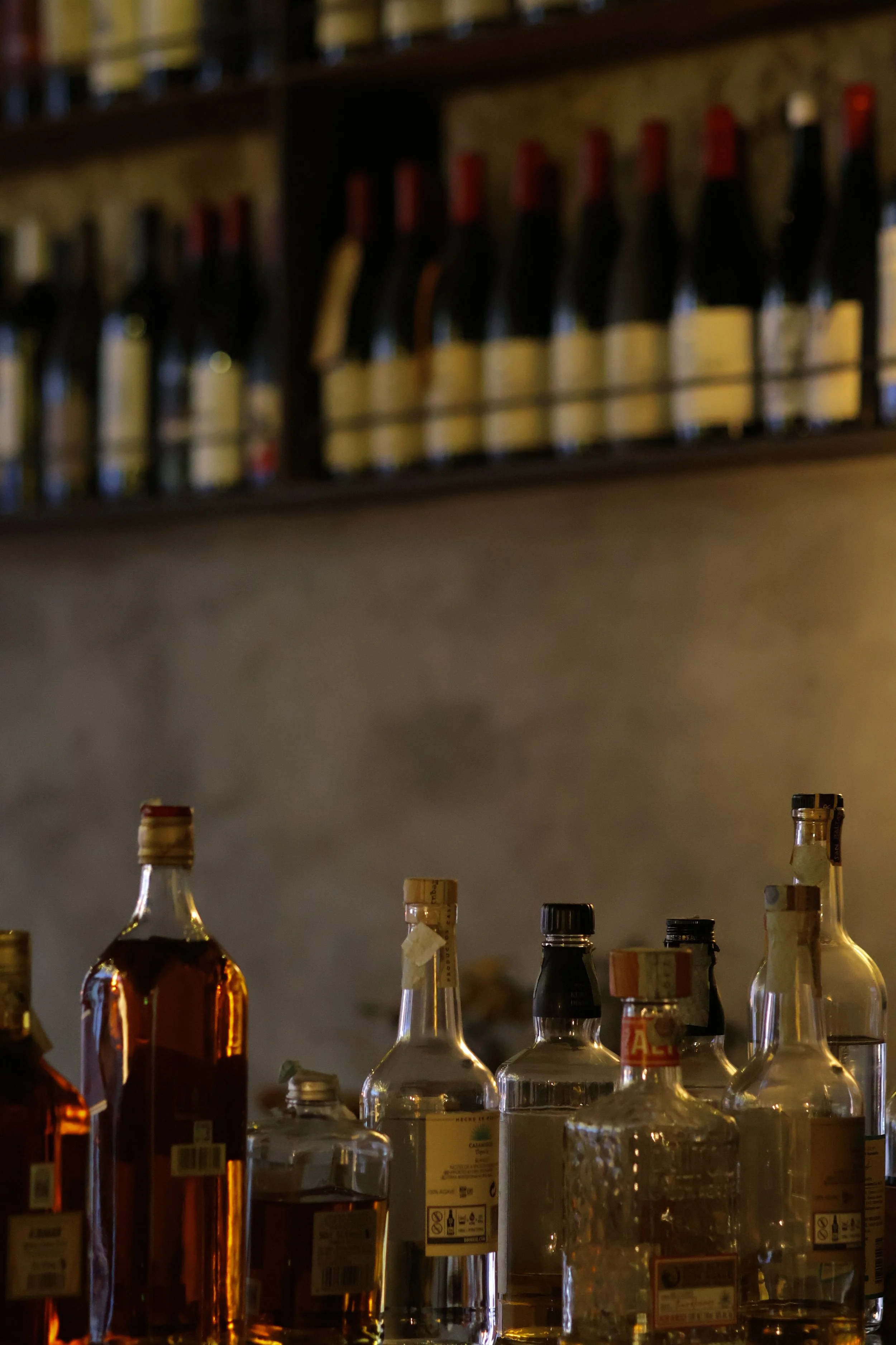 Aesthetic photo in a restaurant of a collection of wine bottles on a bar counter with a  shelf of bottles in the background.