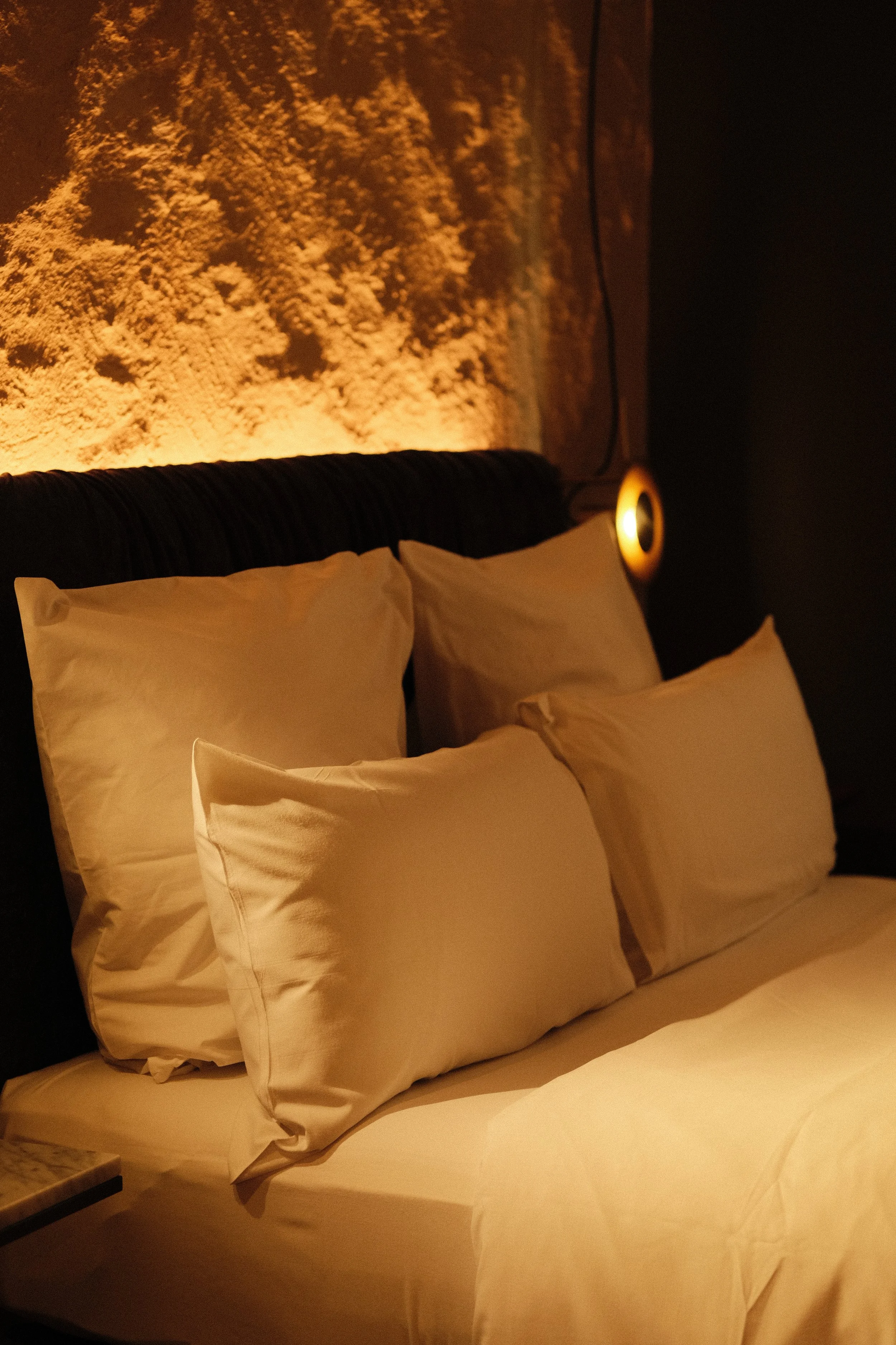 A cozy bedroom with a bed featuring four pillows, a softly glowing wall lamp, and a textured wall art above the bed.