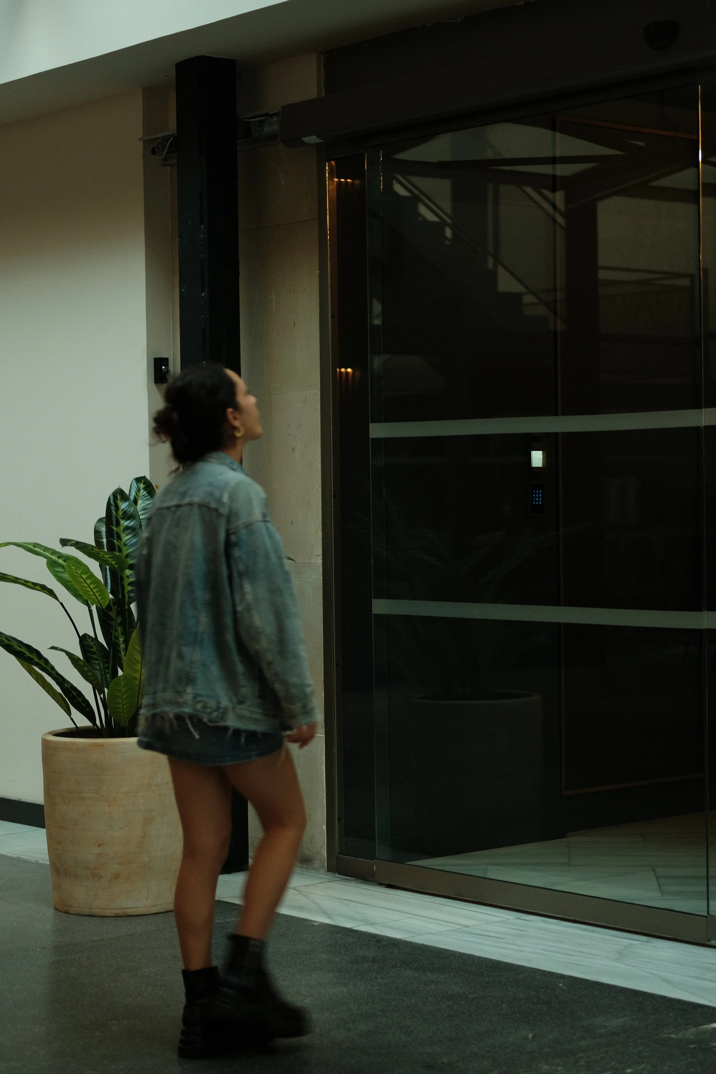 A woman with a denim jacket and black boots standing in front of an elevator with a potted plant nearby.