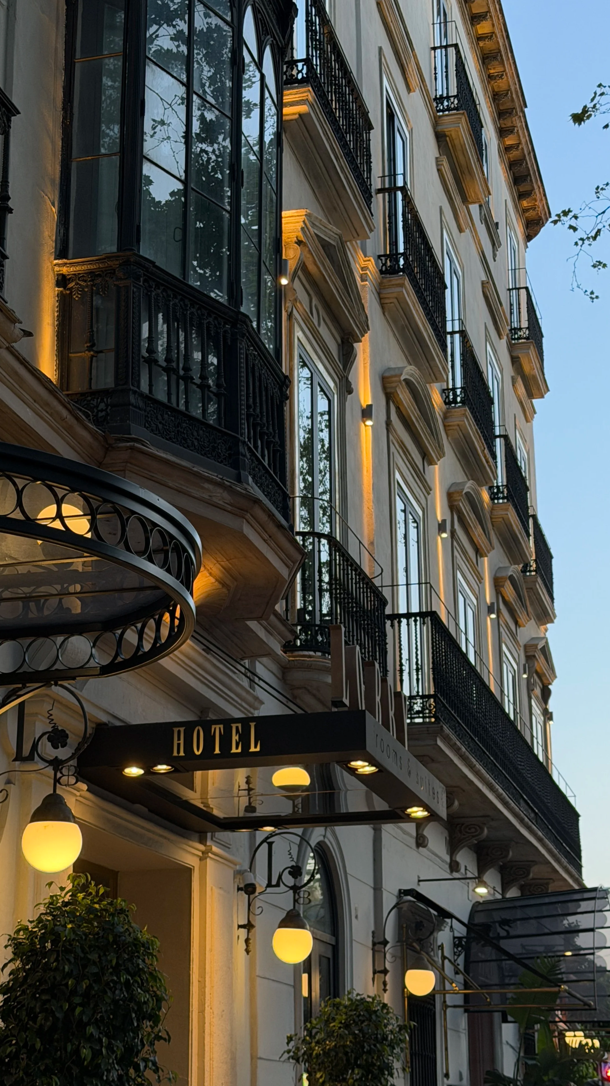 Exterior view of a hotel building during evening with illuminated round lamps, decorative iron balconies, potted plants, and a black awning with gold letters reading 'HOTEL'