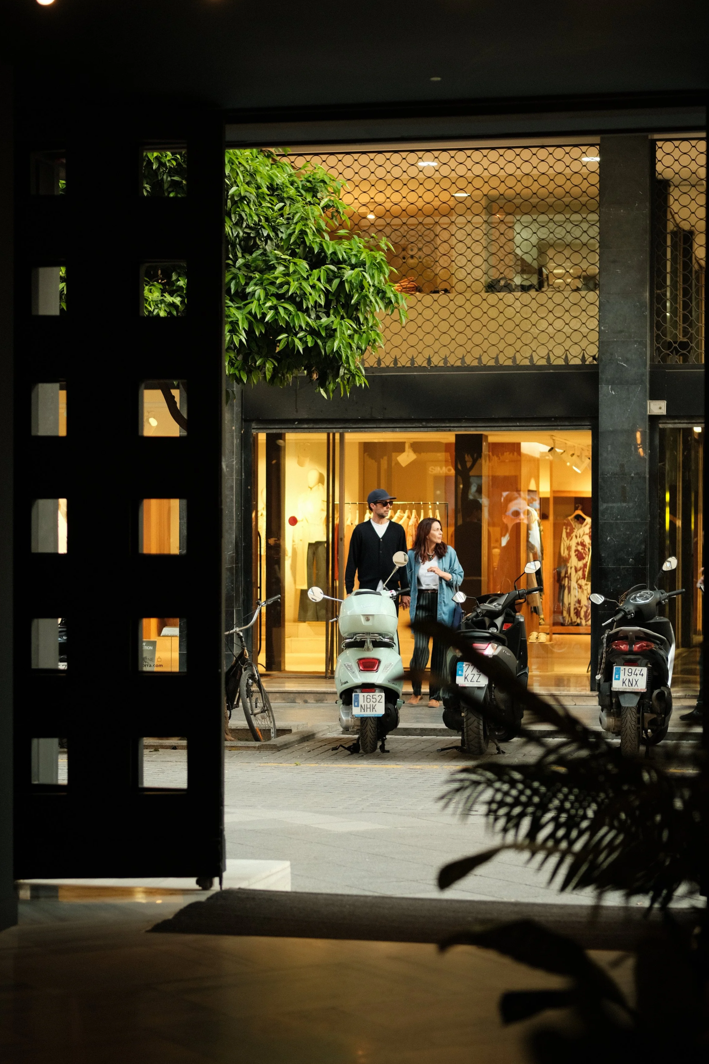 A view from inside a building looking out to a street scene with two people standing in front of a fashion boutique, several parked scooters, and a bicycle, with the boutique’s interior illuminated.