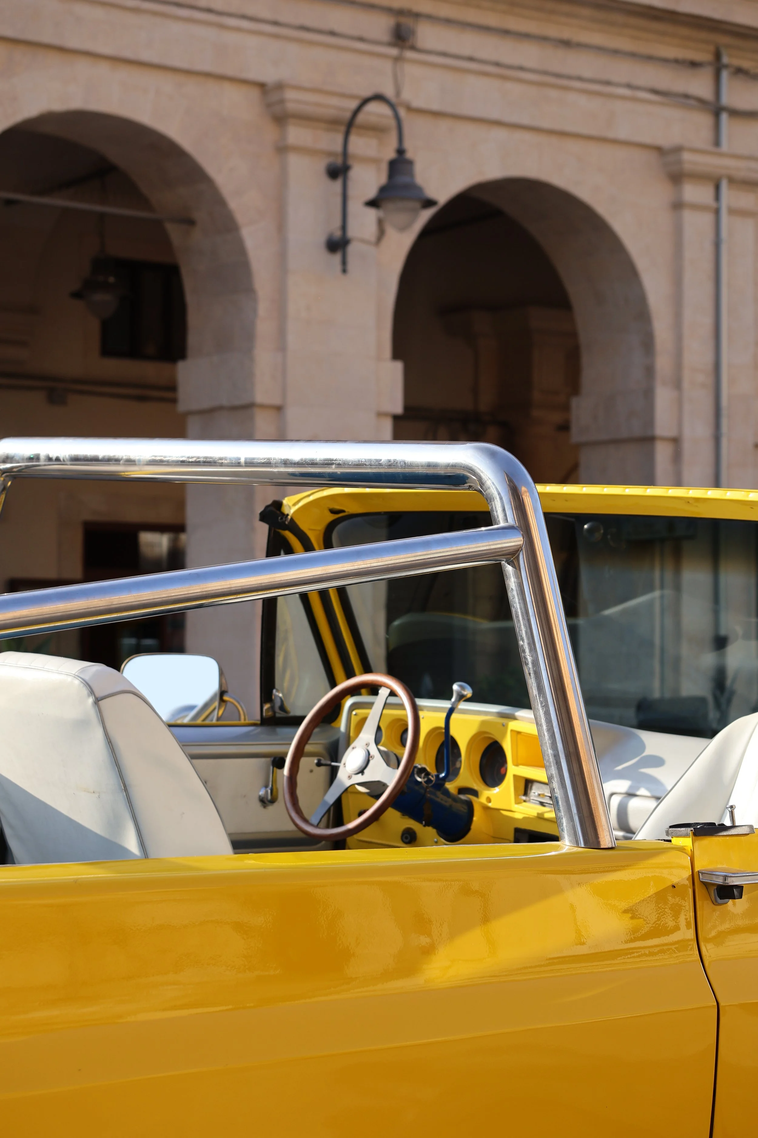 Close-up of a yellow vintage convertible car with white seats, viewed from the side, against a background of a beige stone building with arches and hanging lamps.
