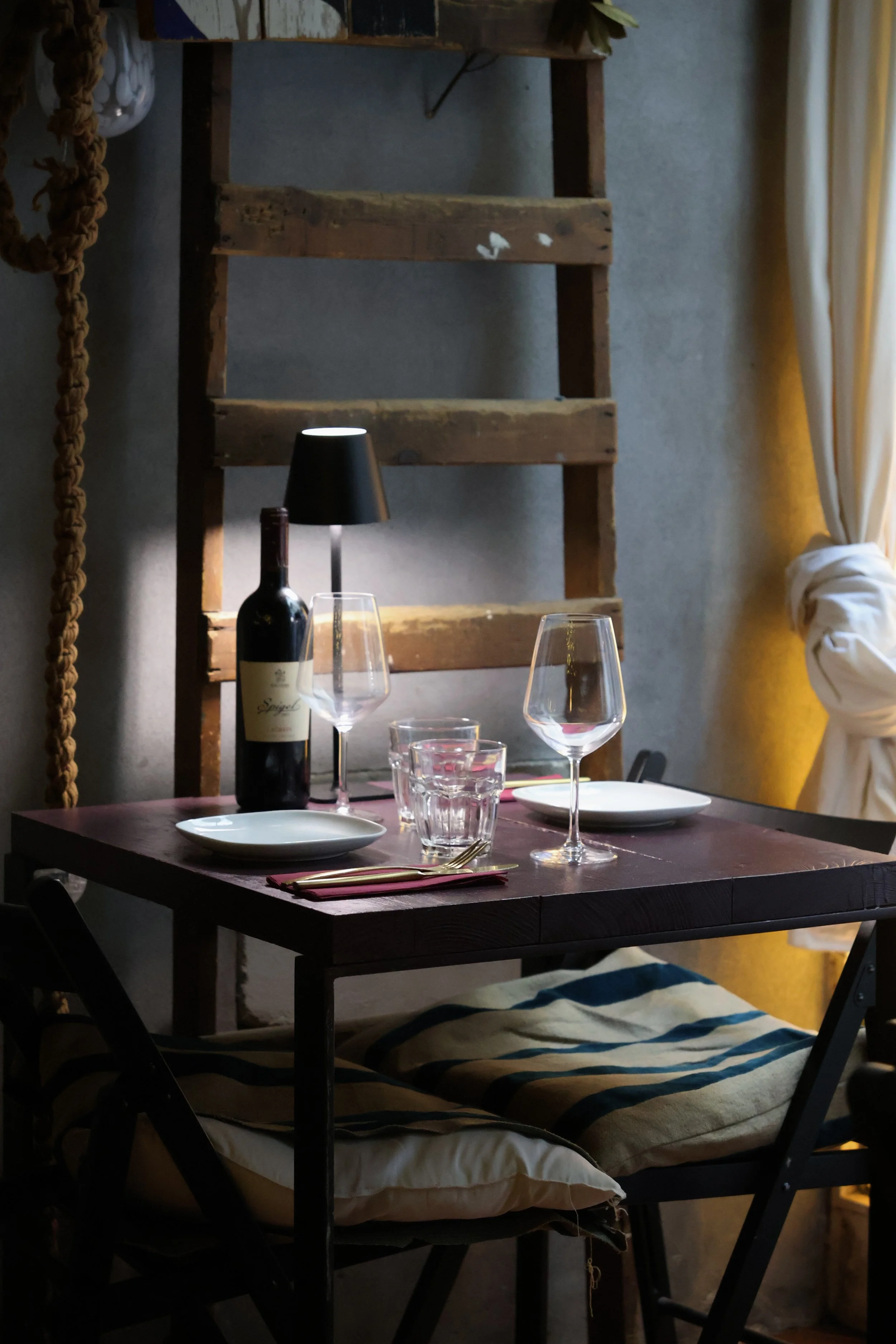A dining table set for three with two empty wine glasses, a bottle of wine, water glasses, white plates, and cutlery. The table is illuminated by a small black table lamp and positioned near a window with white curtains.