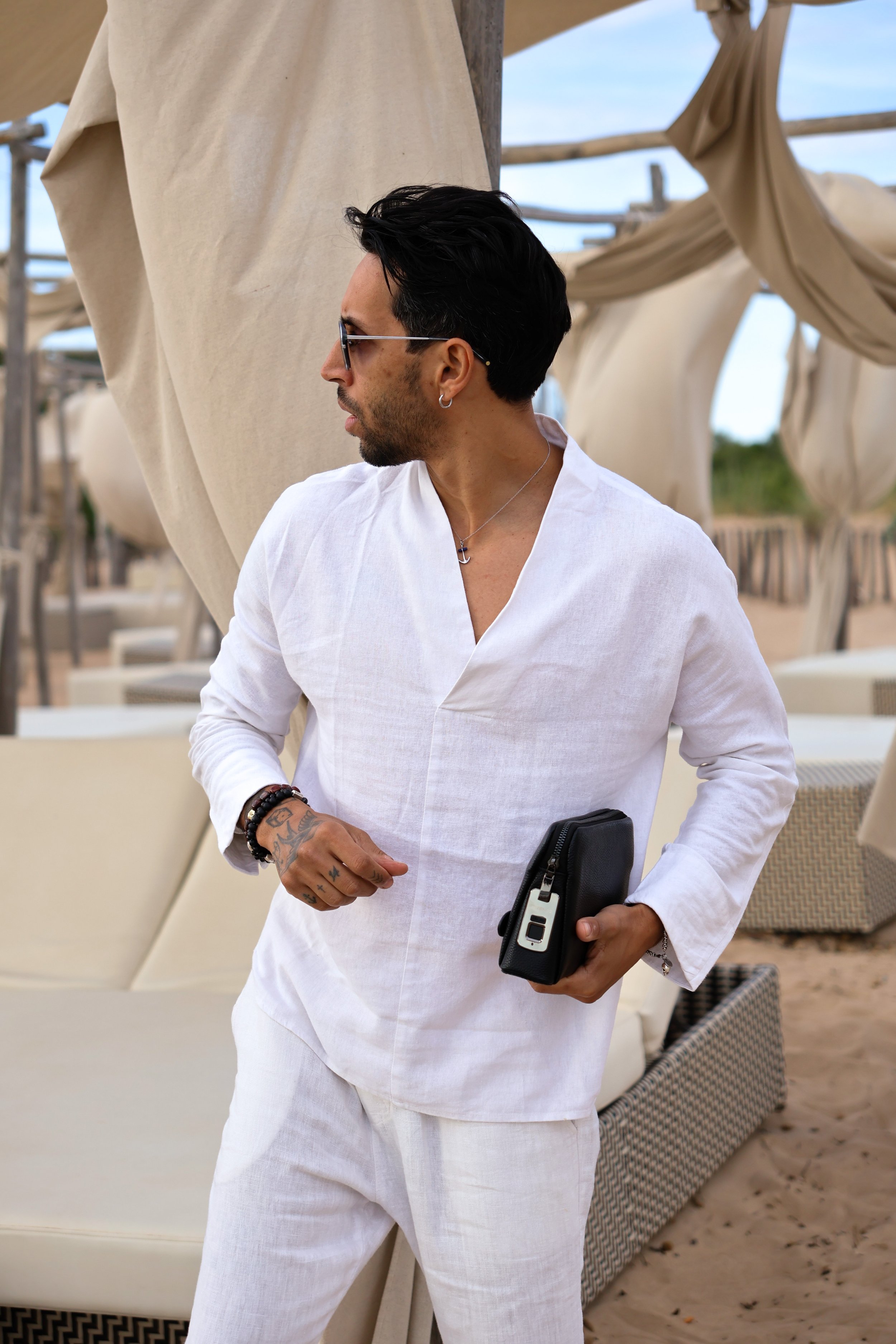 A man dressed in white linen outfit holding a black clutch purse, standing near beach loungers with beige curtains in the background.