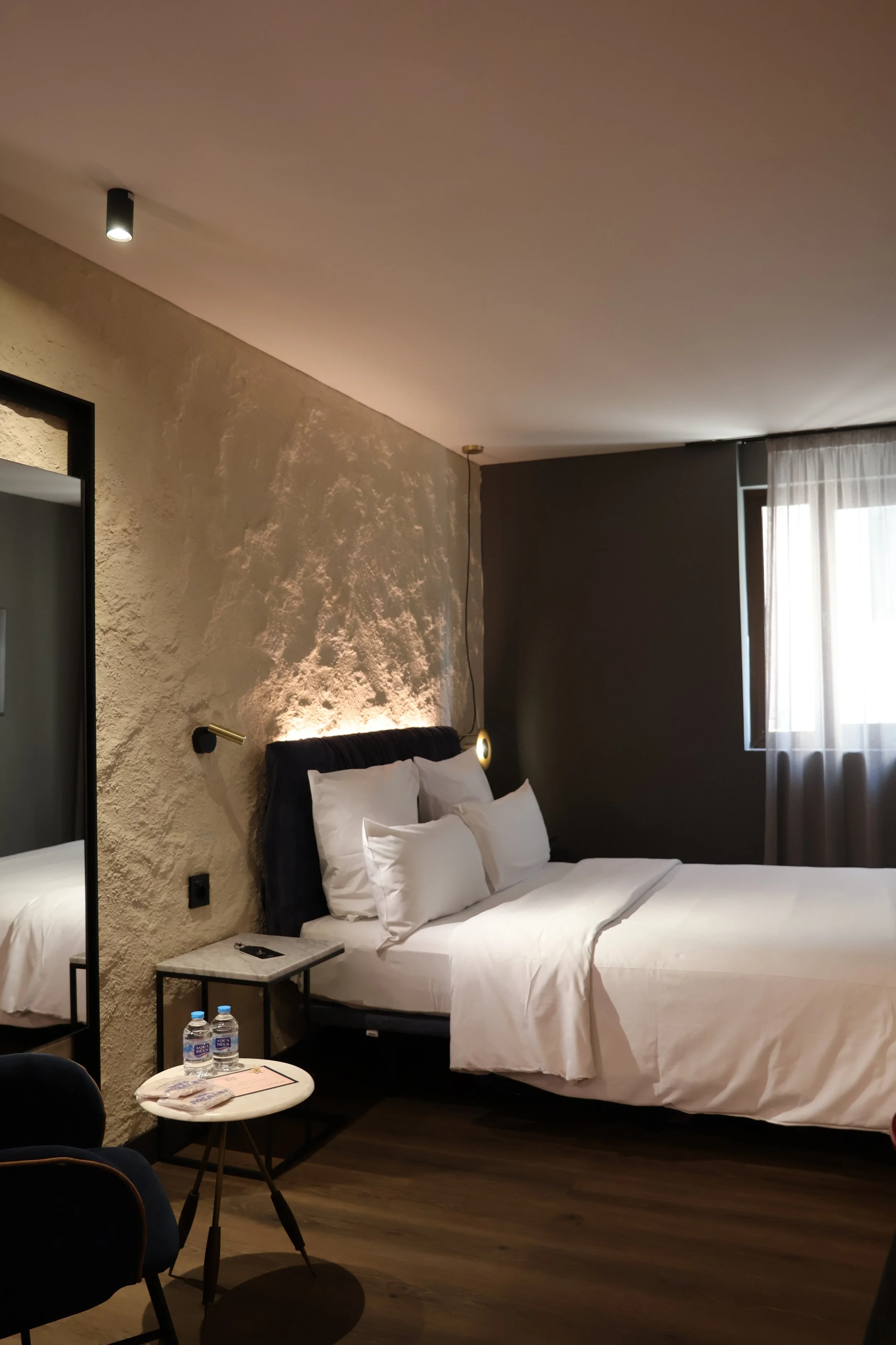 A modern hotel room with a bed, bedside table, two water bottles, a mirror, and a window with sheer curtains.