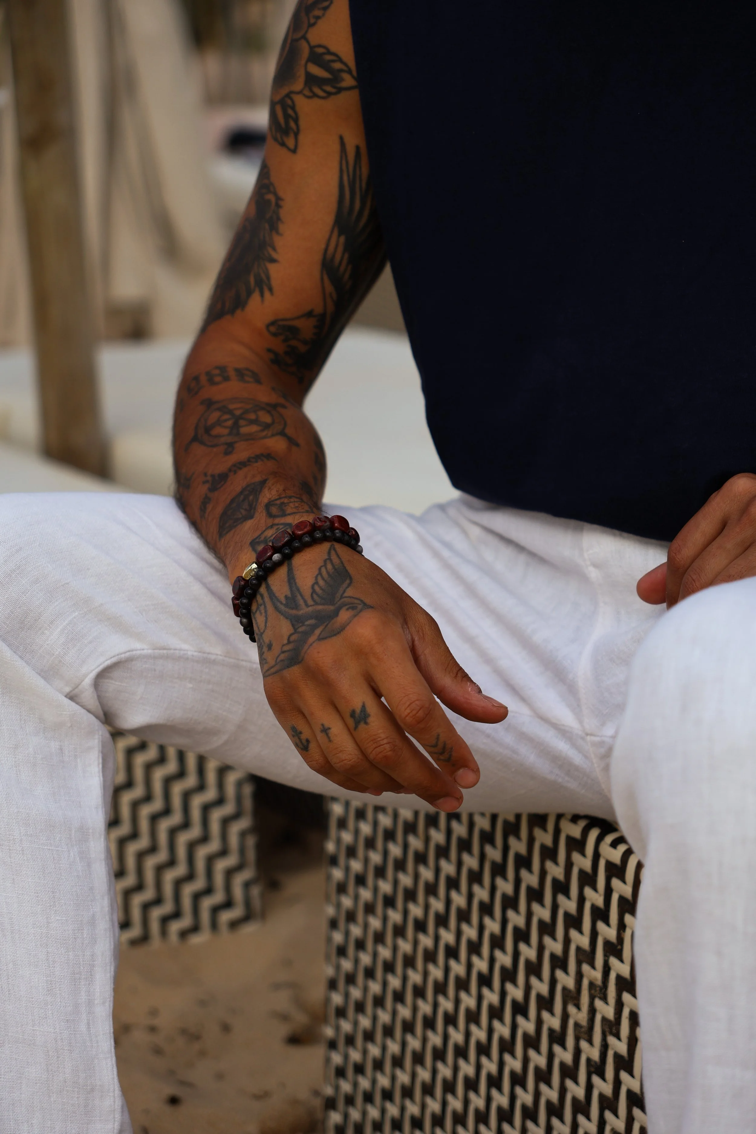 A person sitting on a bench, wearing a navy blue t-shirt and white pants. The person's left arm and hand, which are covered in various tattoos, are resting on their knee. The tattoos include birds, crosses, detailed designs, and text. The individual 