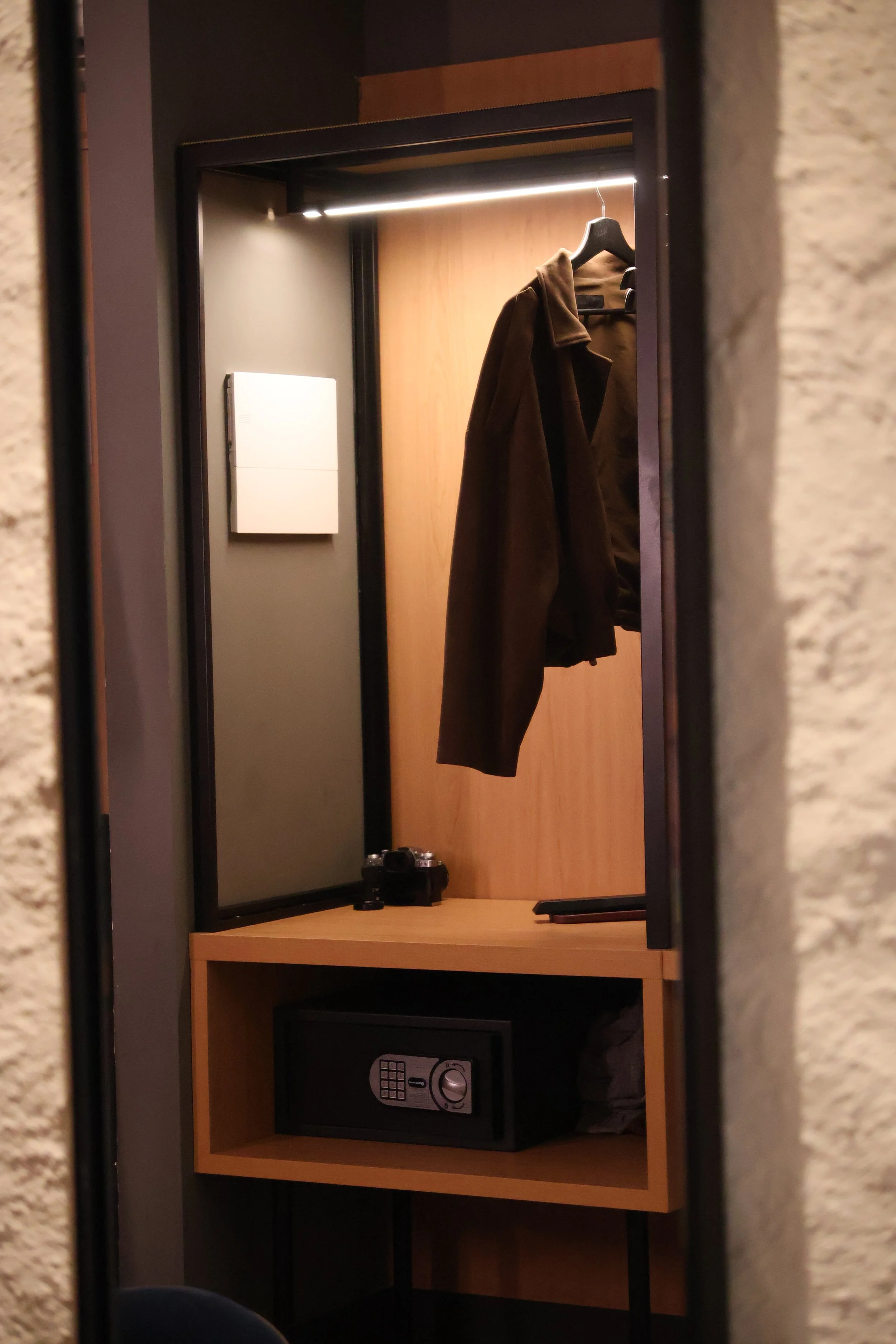 View inside a hotel room closet with brown coat hanging on a hanger, a black camera on the wooden shelf, and a safe in the lower compartment.