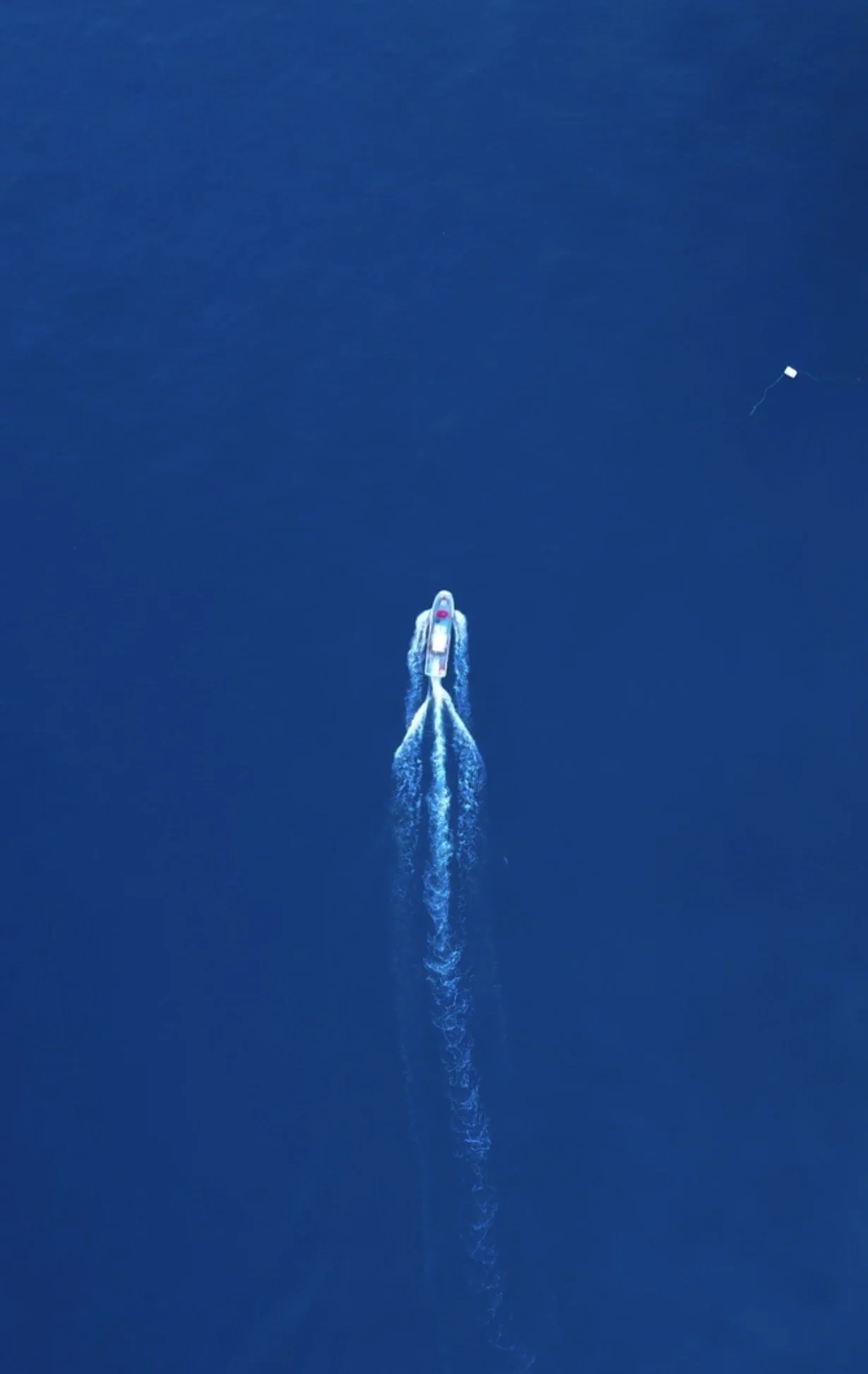 An aerial view of a speedboat creating a white wake on deep blue water with a small white drone in the distance. Drone shot.