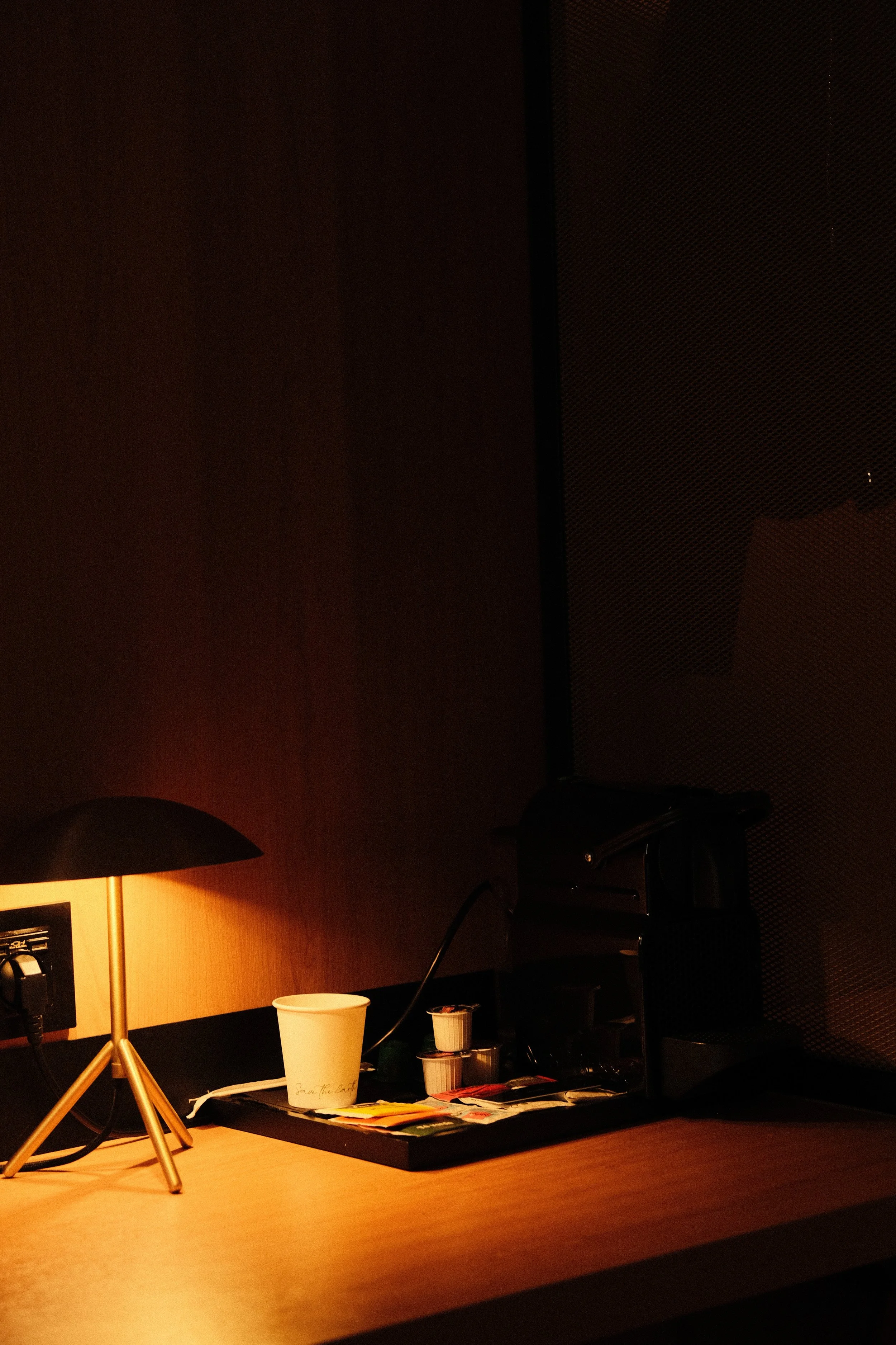 A wooden desk with a small black lamp, a paper cup, sugar containers, and a coffee machine, against a dark wall with textured paneling.