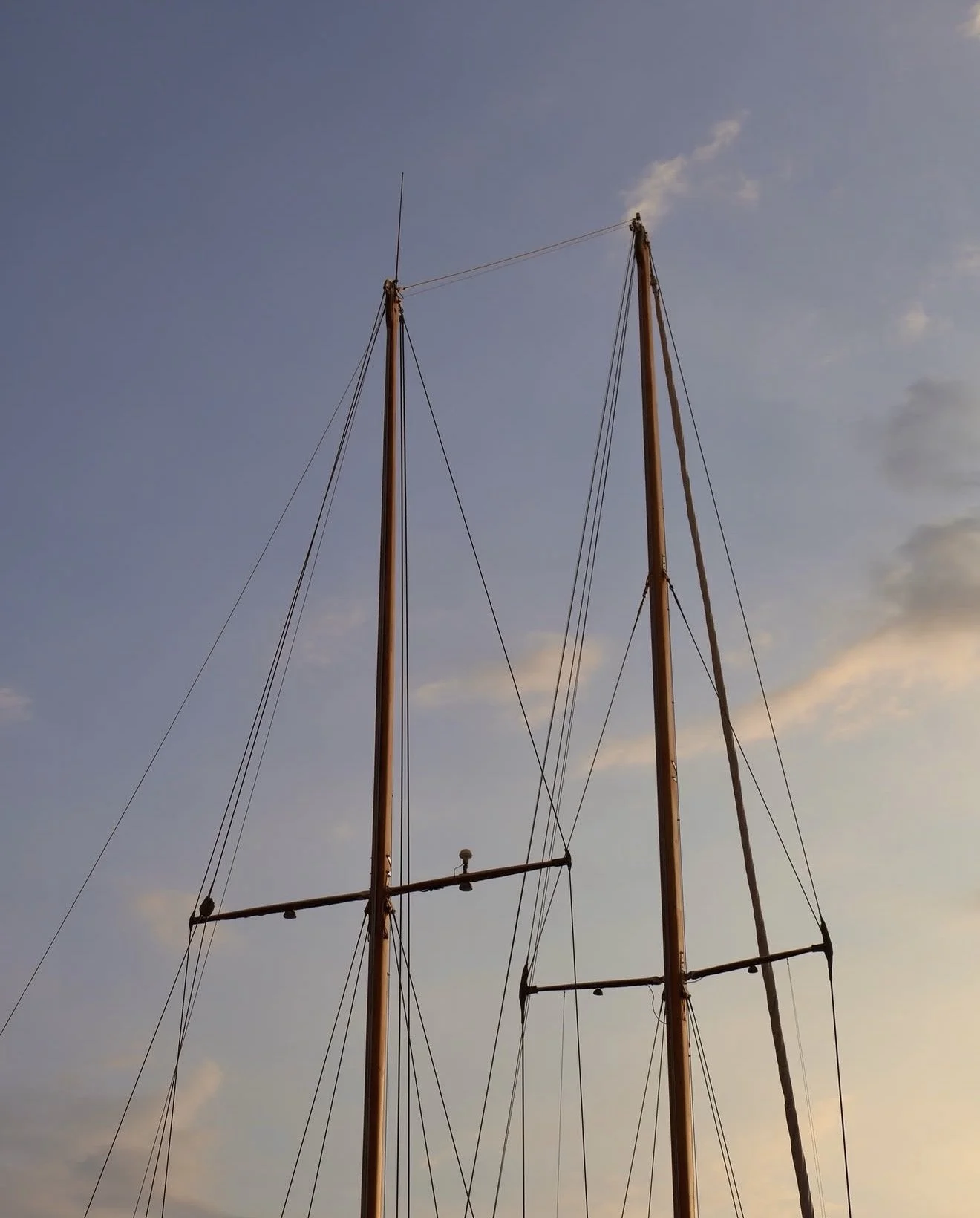 Two wooden boat masts with rigging against a blue sky with some clouds.
