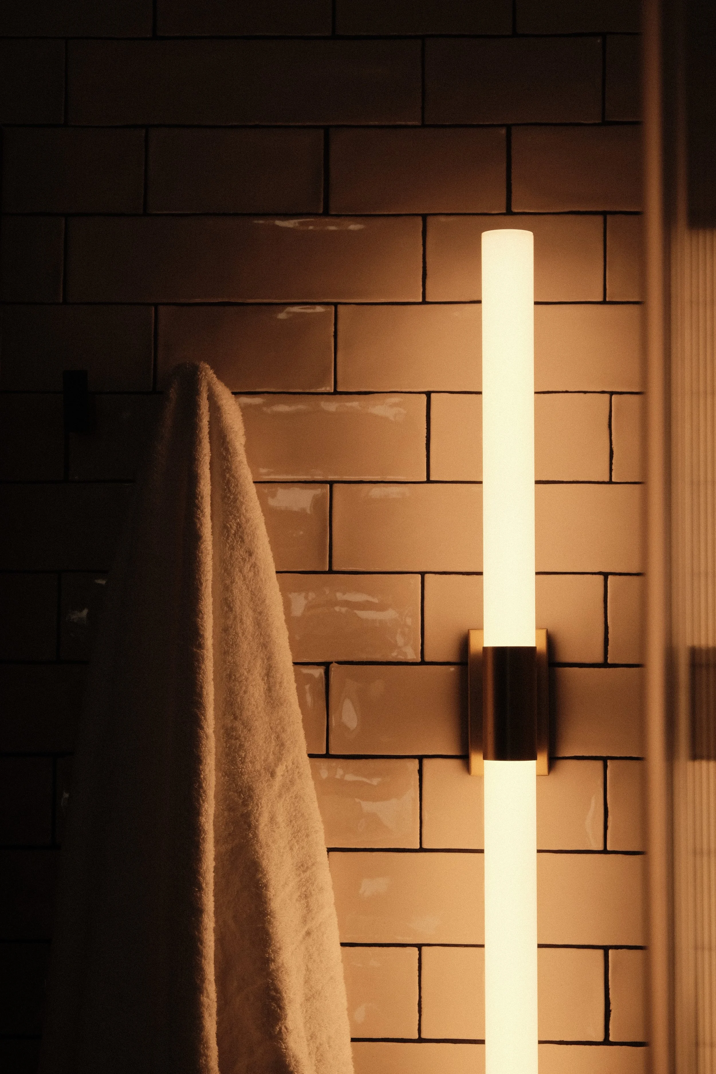 A beige towel hangs on a black hook on a brick wall illuminated by a vertical wall-mounted light fixture with a warm glow.