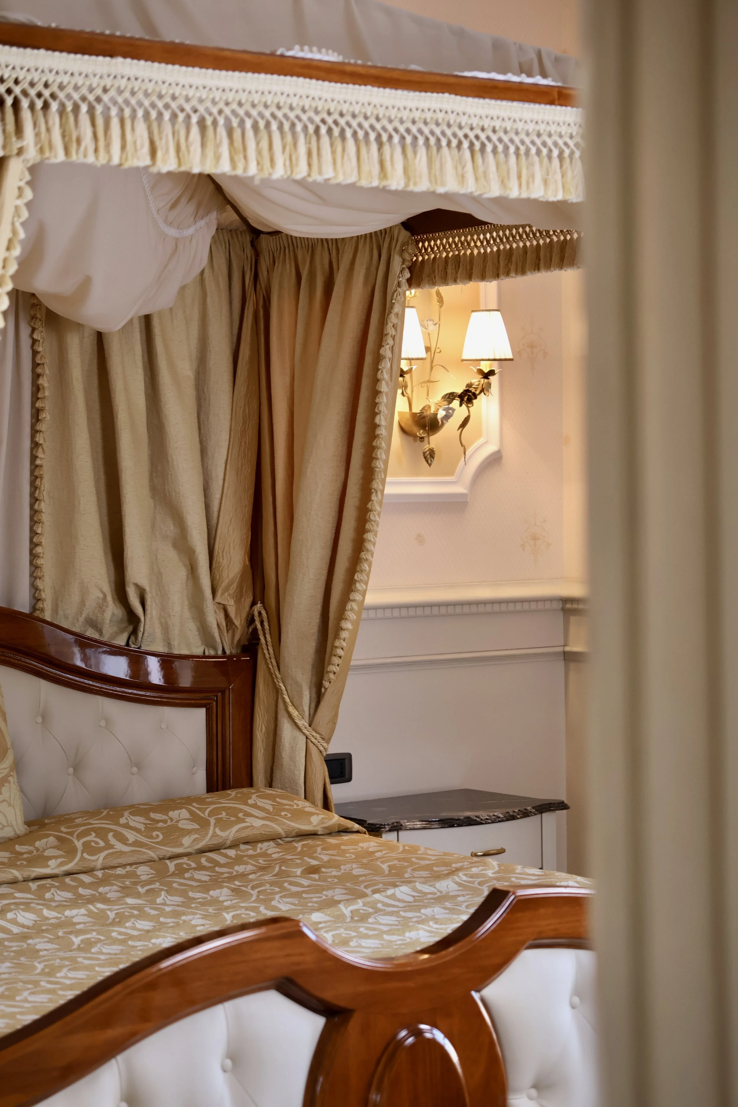 Luxurious bedroom with a four-poster bed, gold patterned bedding, and draped velvet curtains near a wall-mounted lamp with floral decor.