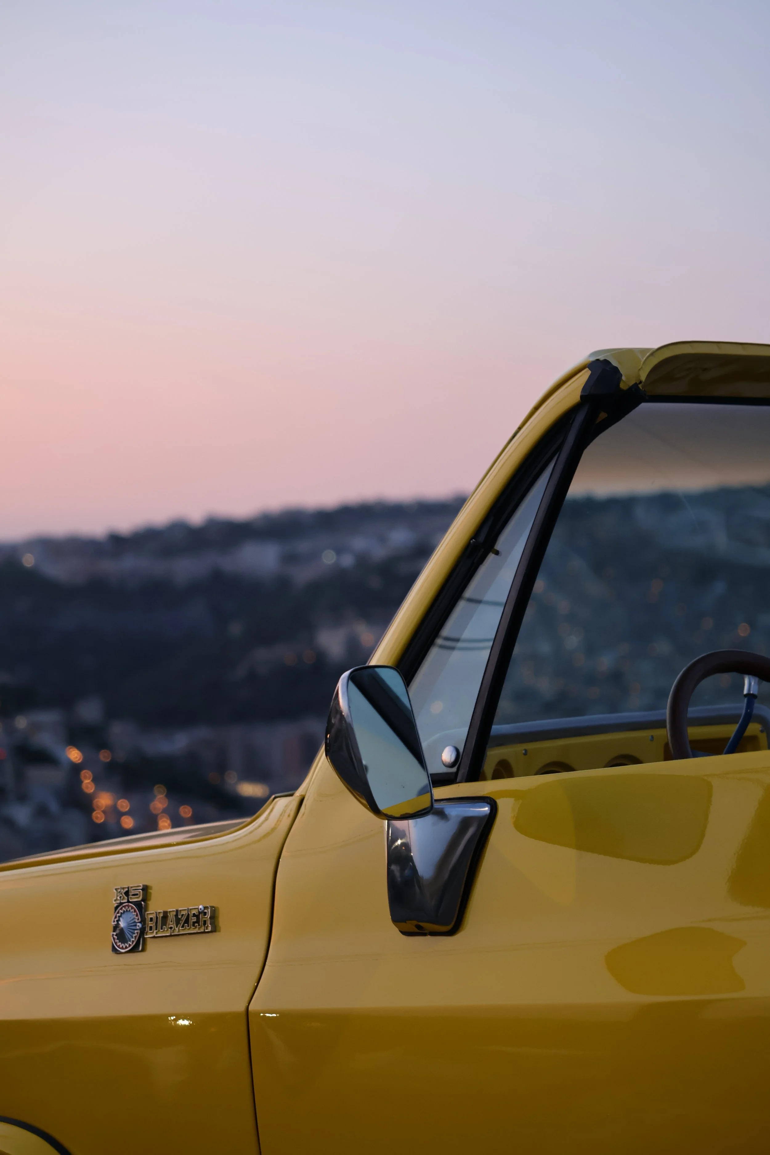Close-up of the front part of a vintage yellow Chevrolet K5 Blazer SUV, showing the side mirror, logo badge, window, and part of the windshield, with a blurred cityscape and hills in the background during dusk.