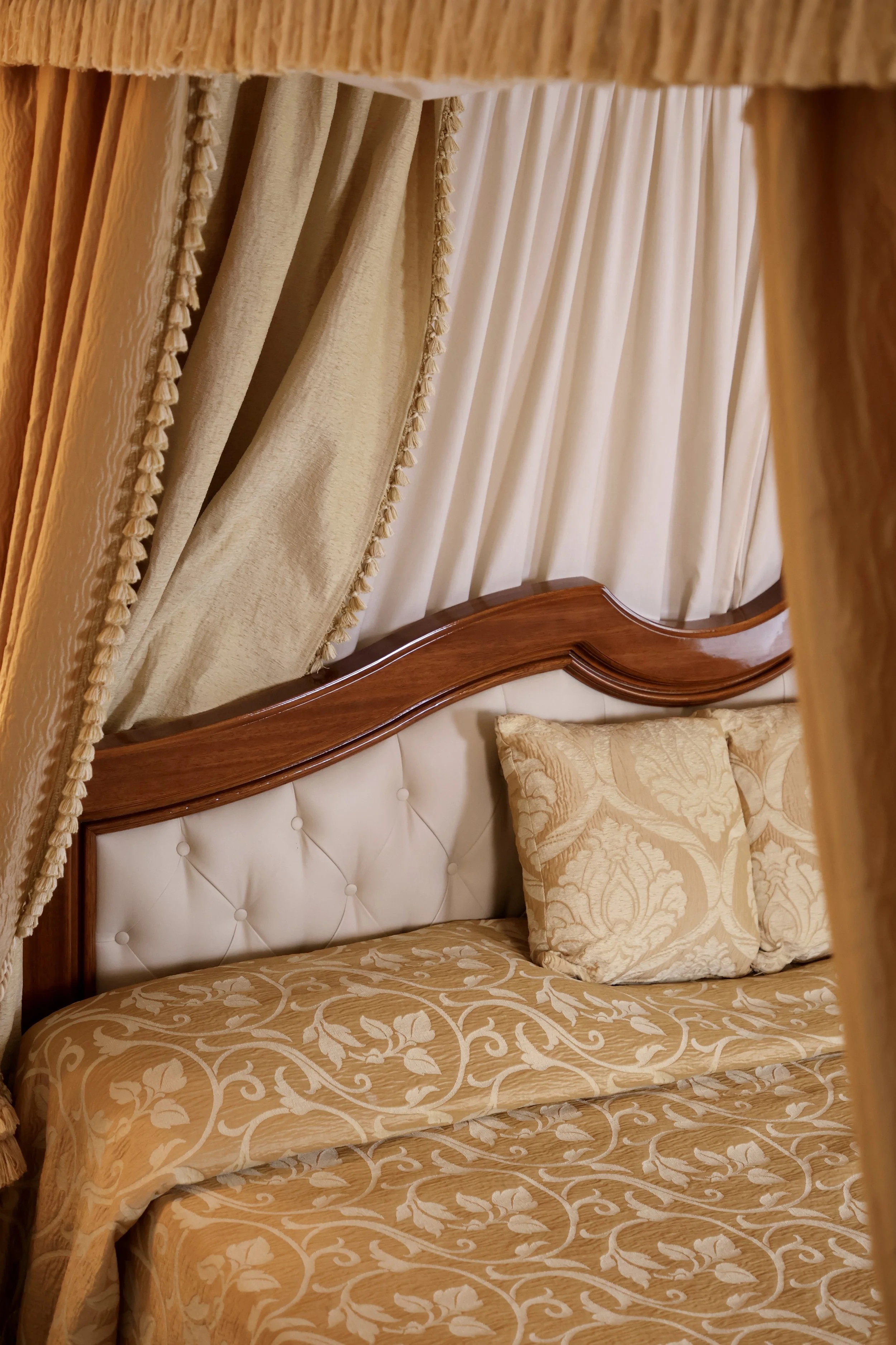 A close-up of a luxurious bed with a wooden headboard, tufted cream leather panel, gold-patterned bedding, and matching pillows, framed by beige curtains.