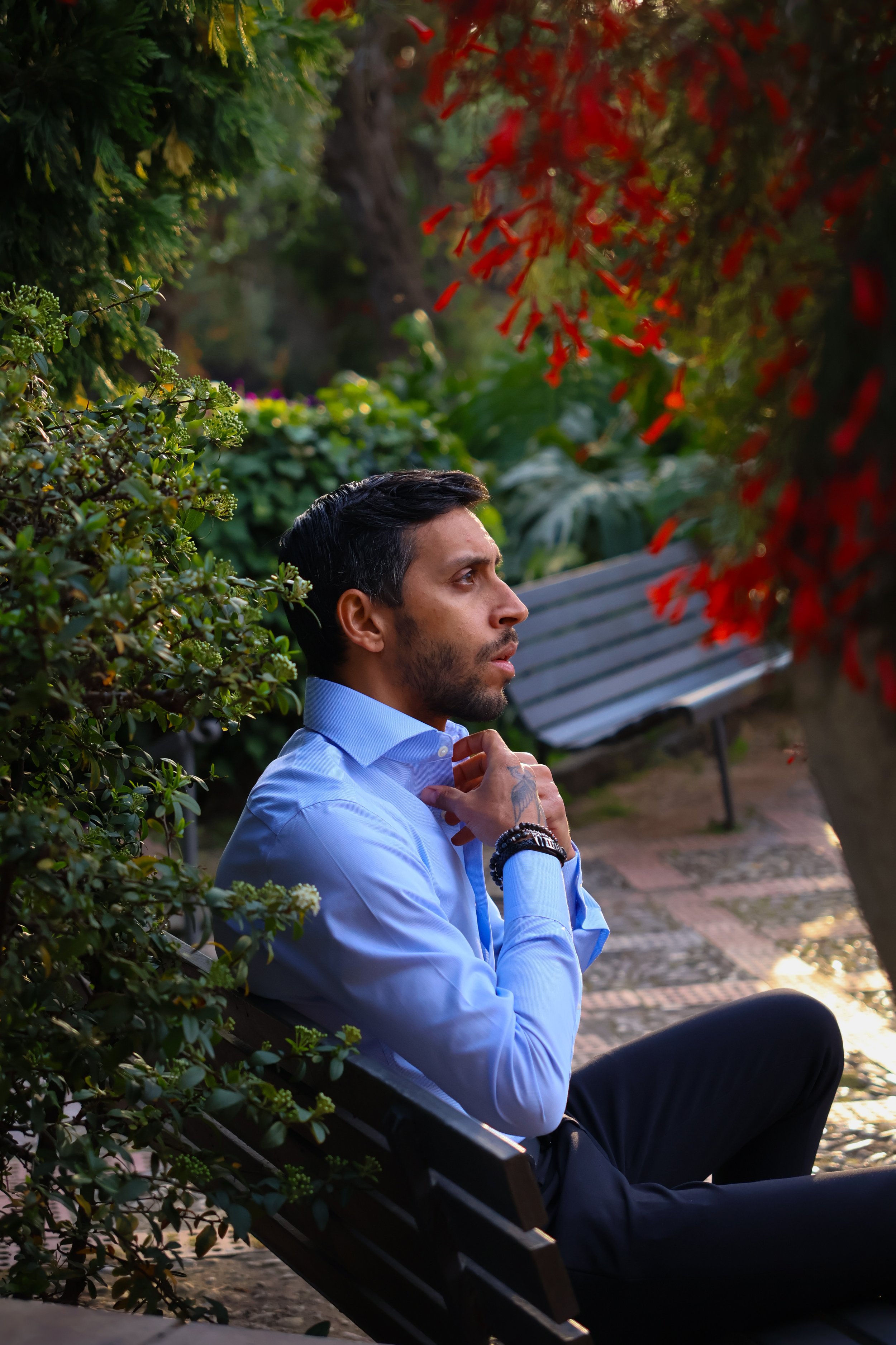 A man in a light blue shirt sitting on a park bench surrounded by greenery and red flowers, appearing to be deep in thought.