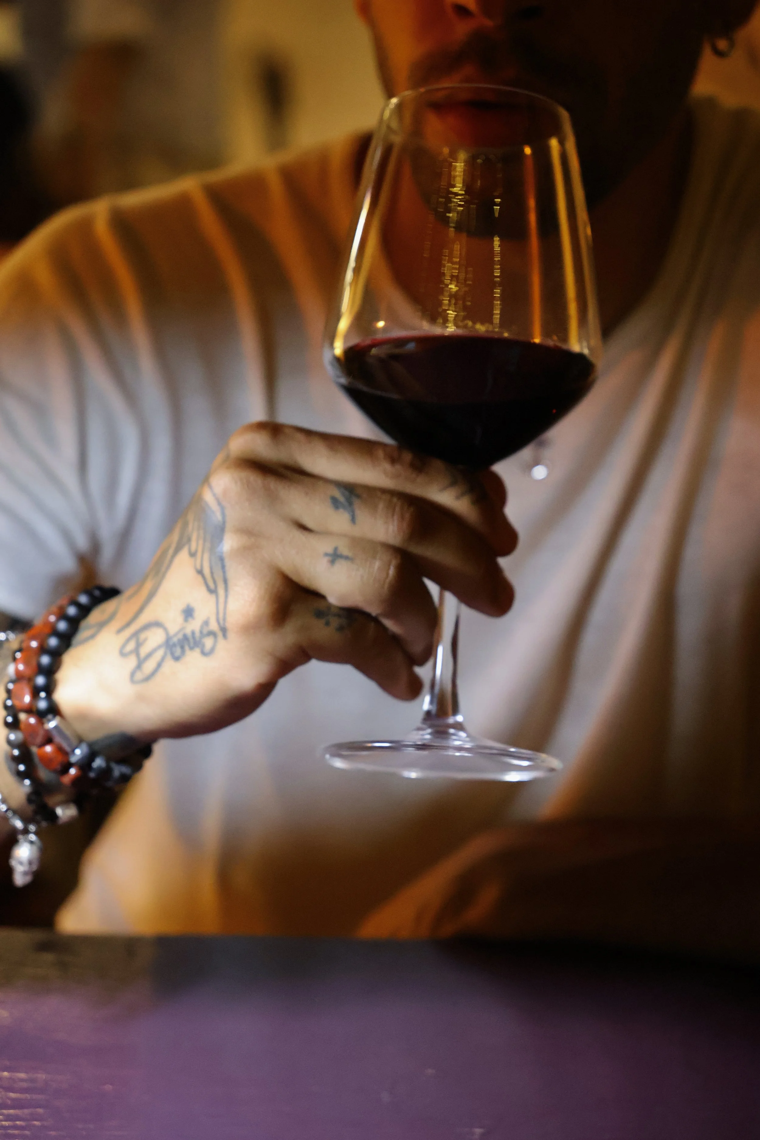 Person holding a glass of red wine, with tattoos and beaded bracelets on their wrist, wearing a light-colored striped shirt, in a dimly lit setting.