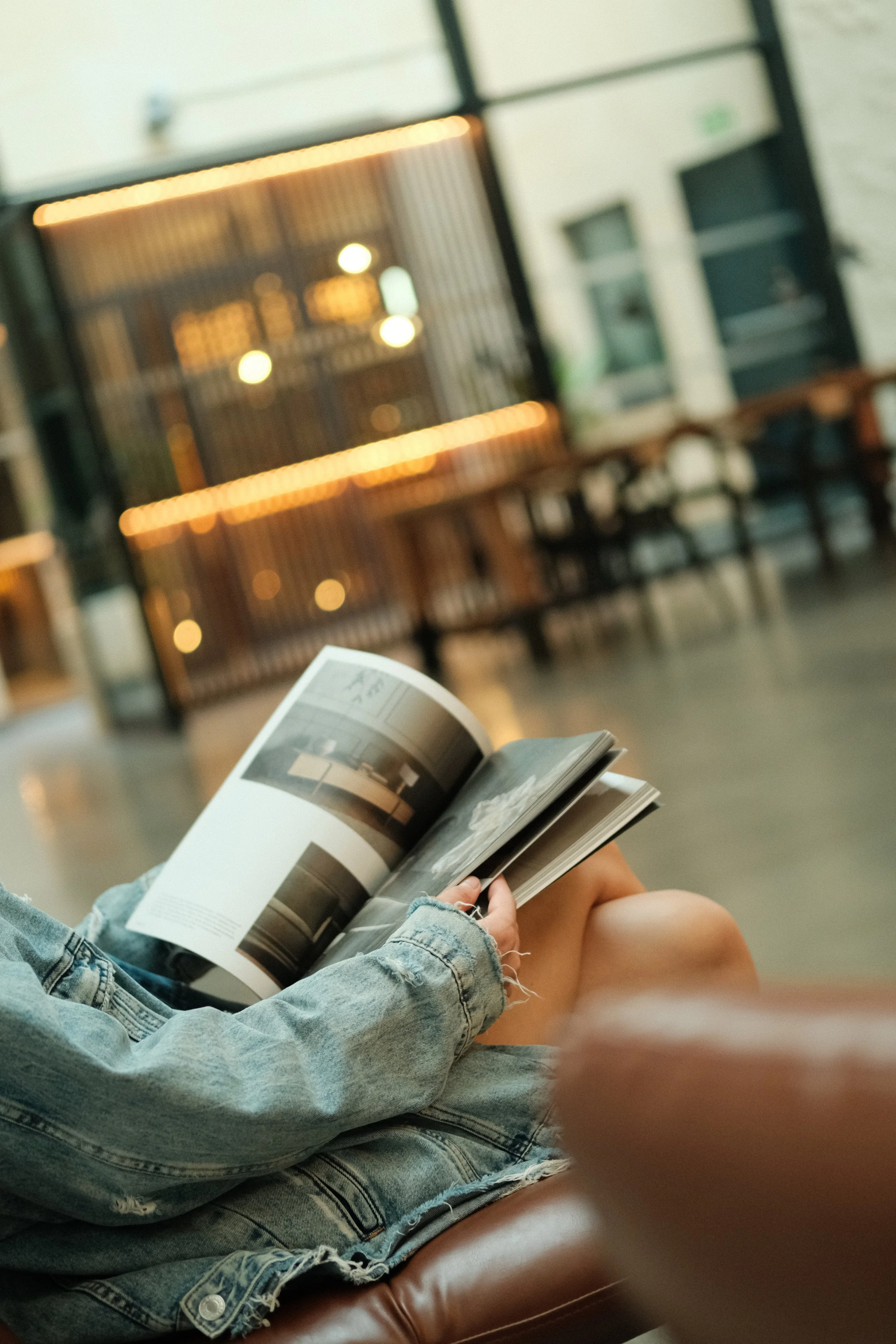 Person in a denim jacket sitting on a leather couch, reading a magazine or book, in a modern indoor space with wooden tables and blurred lighting in the background.