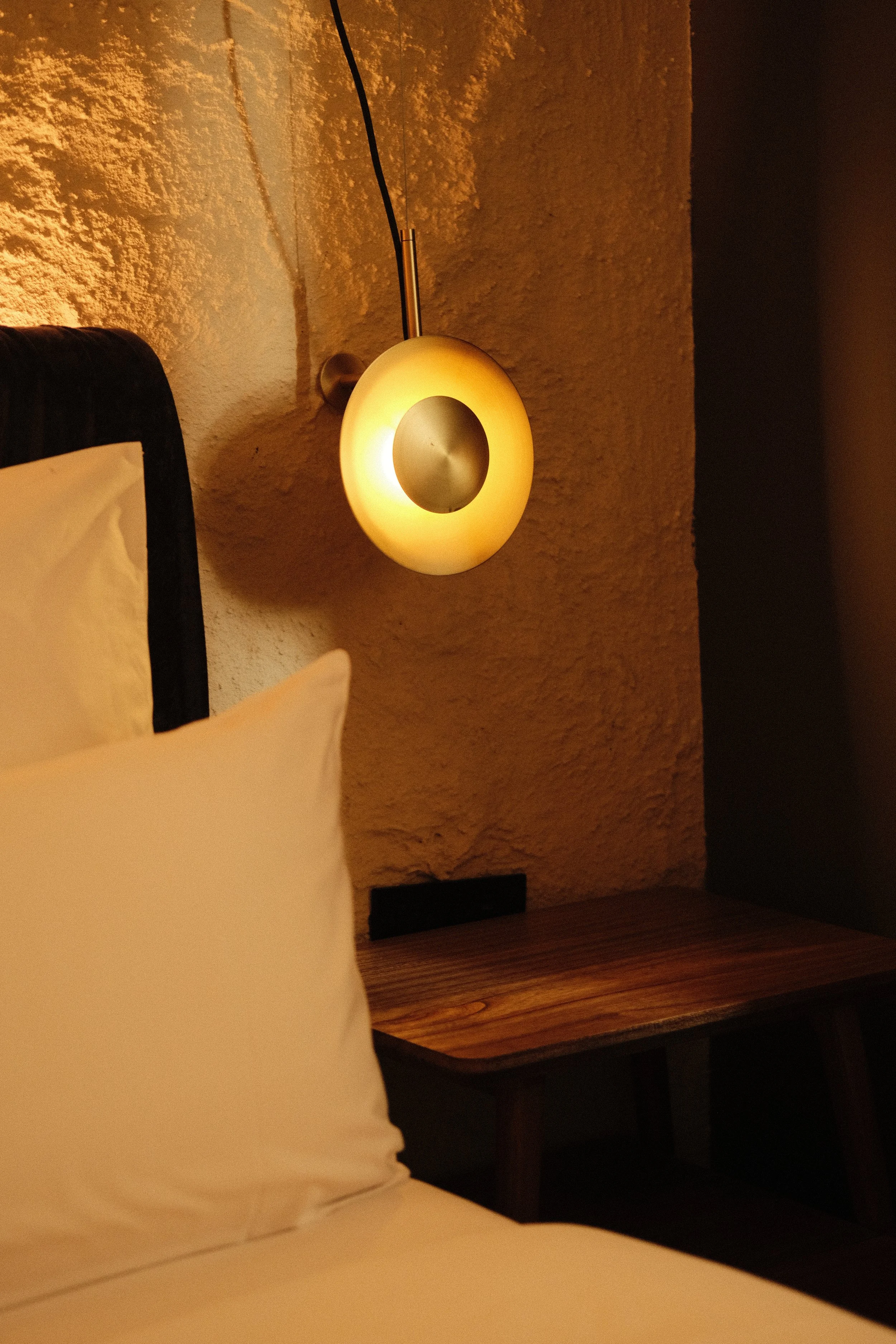 A small round wall-mounted lamp emitting warm yellow light, positioned above a wooden bedside table next to a bed with white pillows, against a textured beige wall.