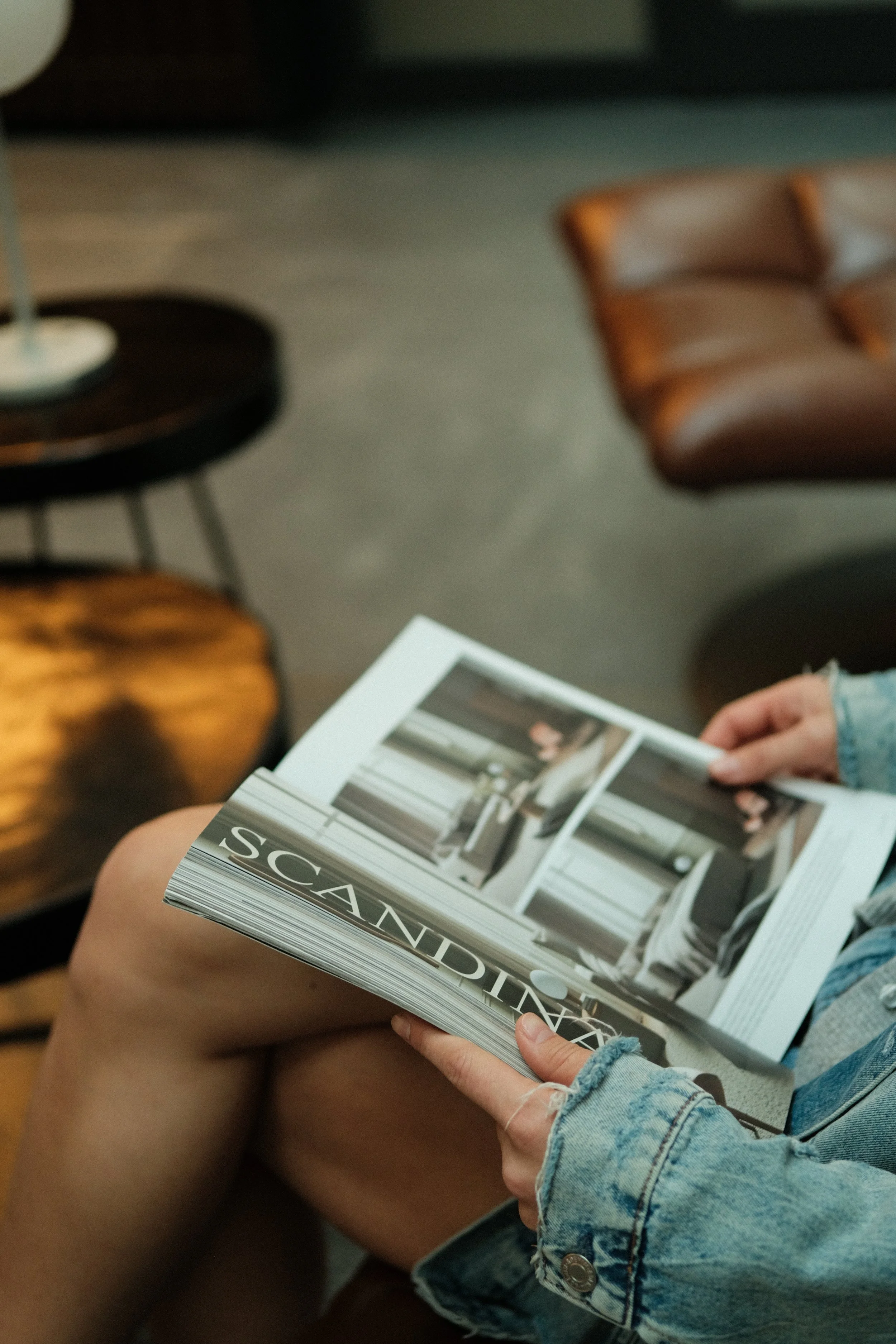 Person in denim jacket holding a magazine with interior design photos, sitting on a chair in a modern living space.