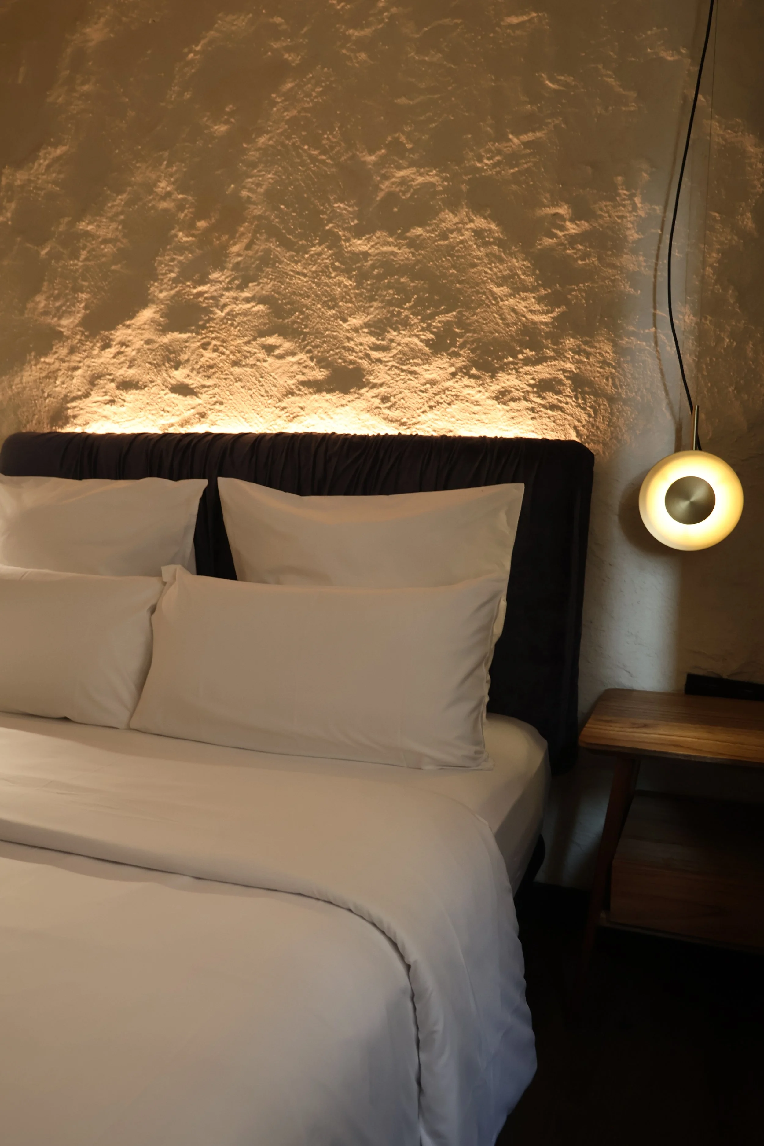 A neatly made bed with white pillows and bed linens, a dark upholstered headboard, a textured beige wall with subtle lighting, and a wooden nightstand with a modern hanging lamp in a cozy bedroom.