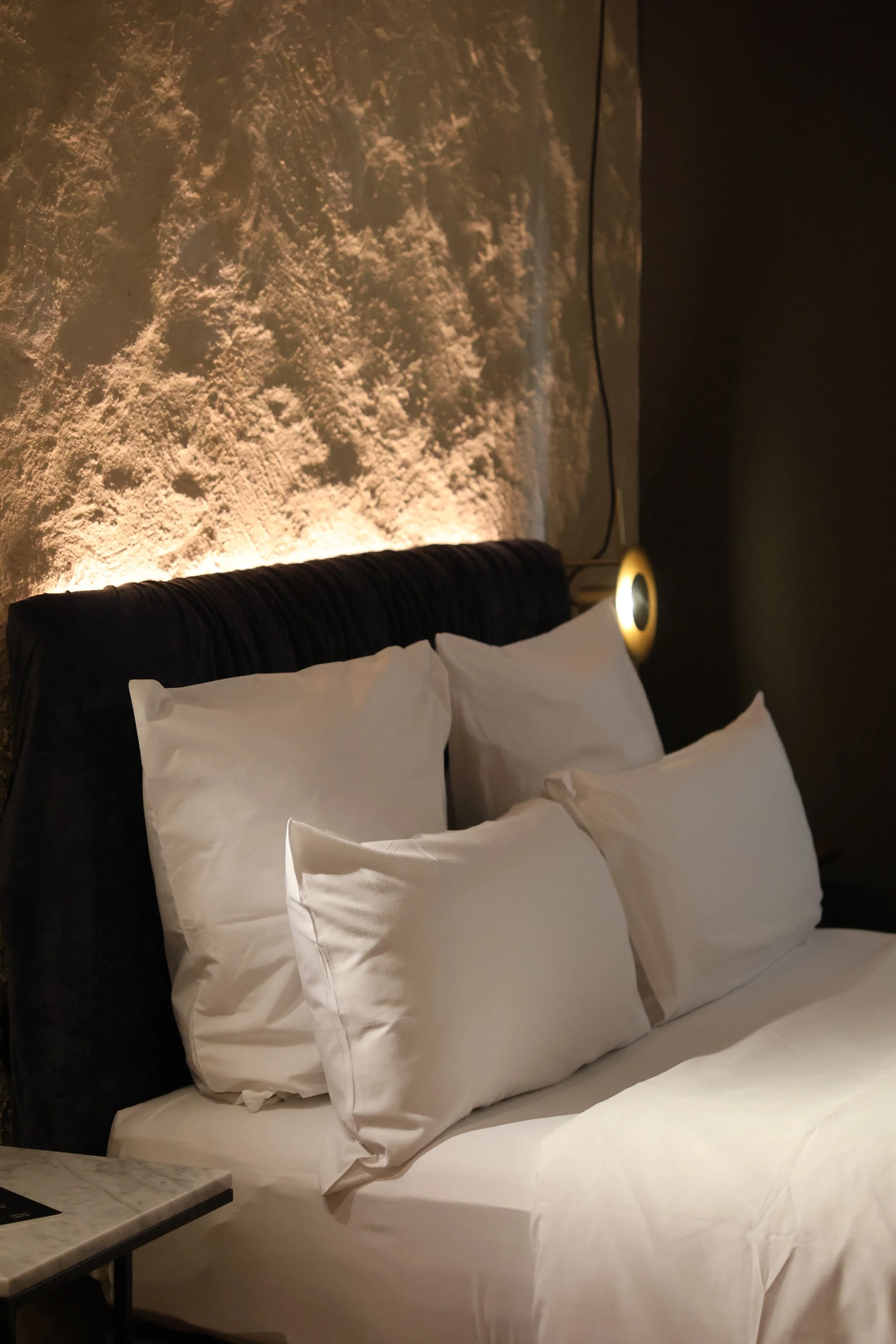 A bed with white pillows and bedding, black headboard, marble side table, textured wall with warm lighting, and a circular wall light fixture.