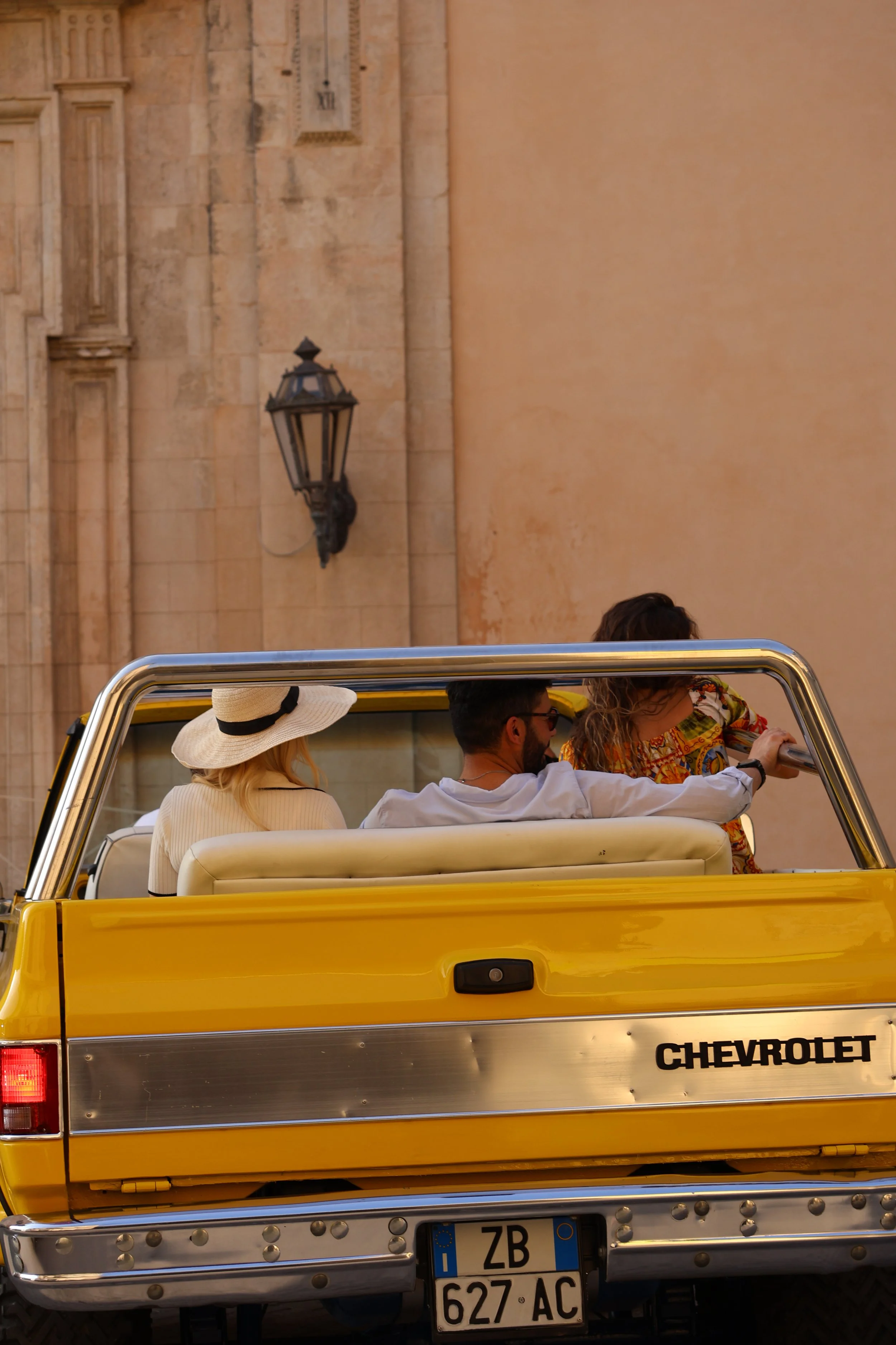 Three people sitting in the back of a yellow Chevrolet pickup truck, with one woman wearing a large sun hat, a man with sunglasses, and a woman with curly hair; the truck is on a street with old building walls and a lantern on the wall.