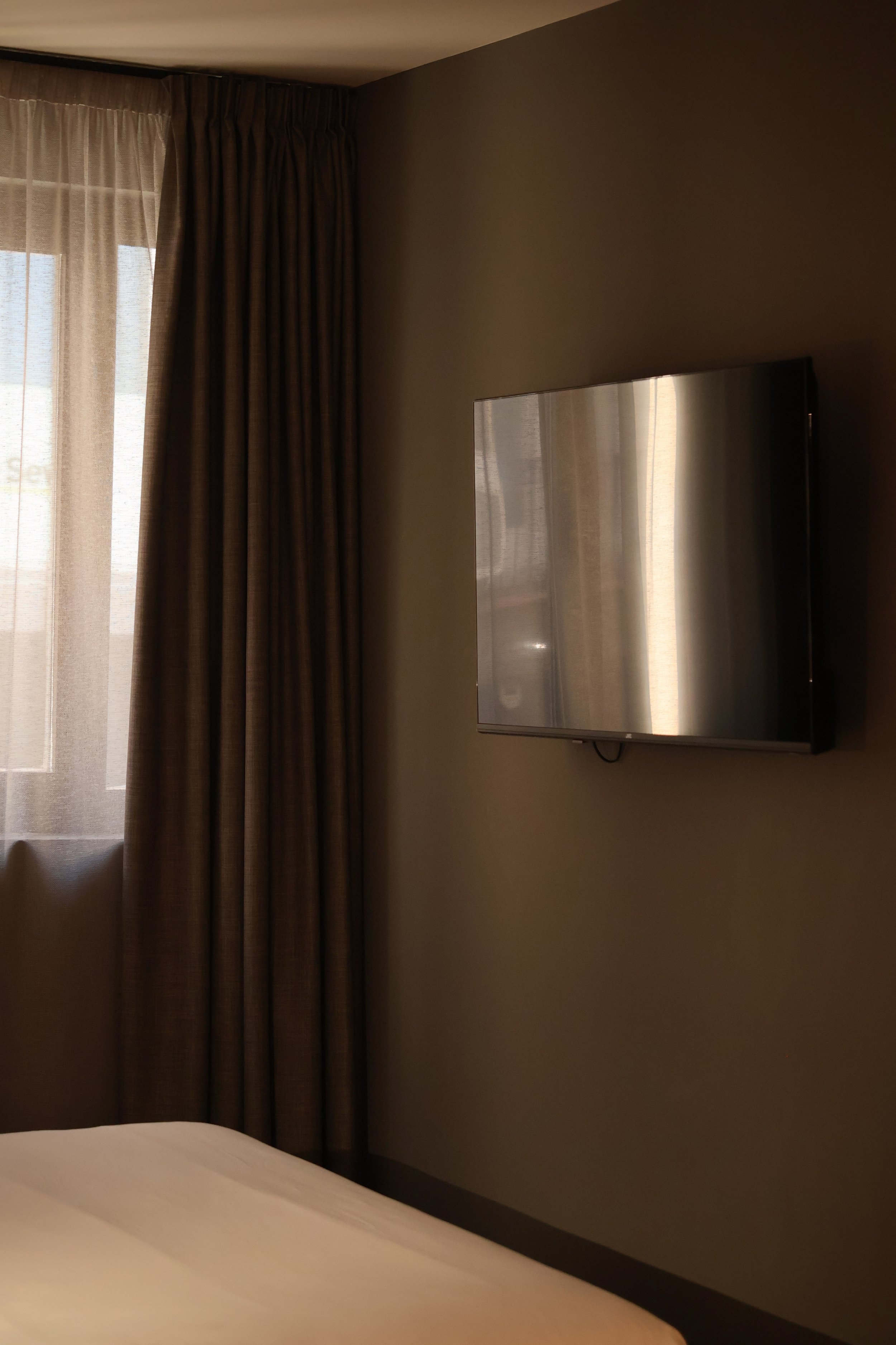 A hotel room featuring a wall-mounted flat-screen TV, a window with sheer and blackout curtains, and part of a bed with white linens.