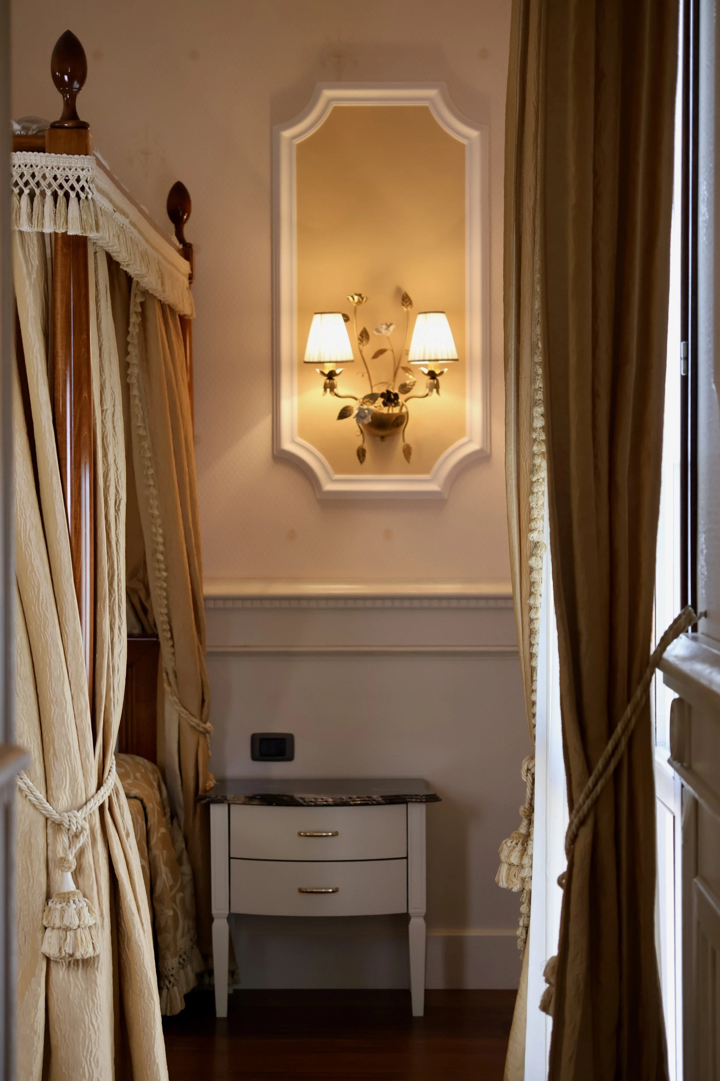 A cozy corner with golden curtains tied back, a small white nightstand with two drawers topped with dark marble, a wall with intricate moulding, a decorative wall sconce with two lamps and floral accents, and part of a four-poster bed with tasseled d