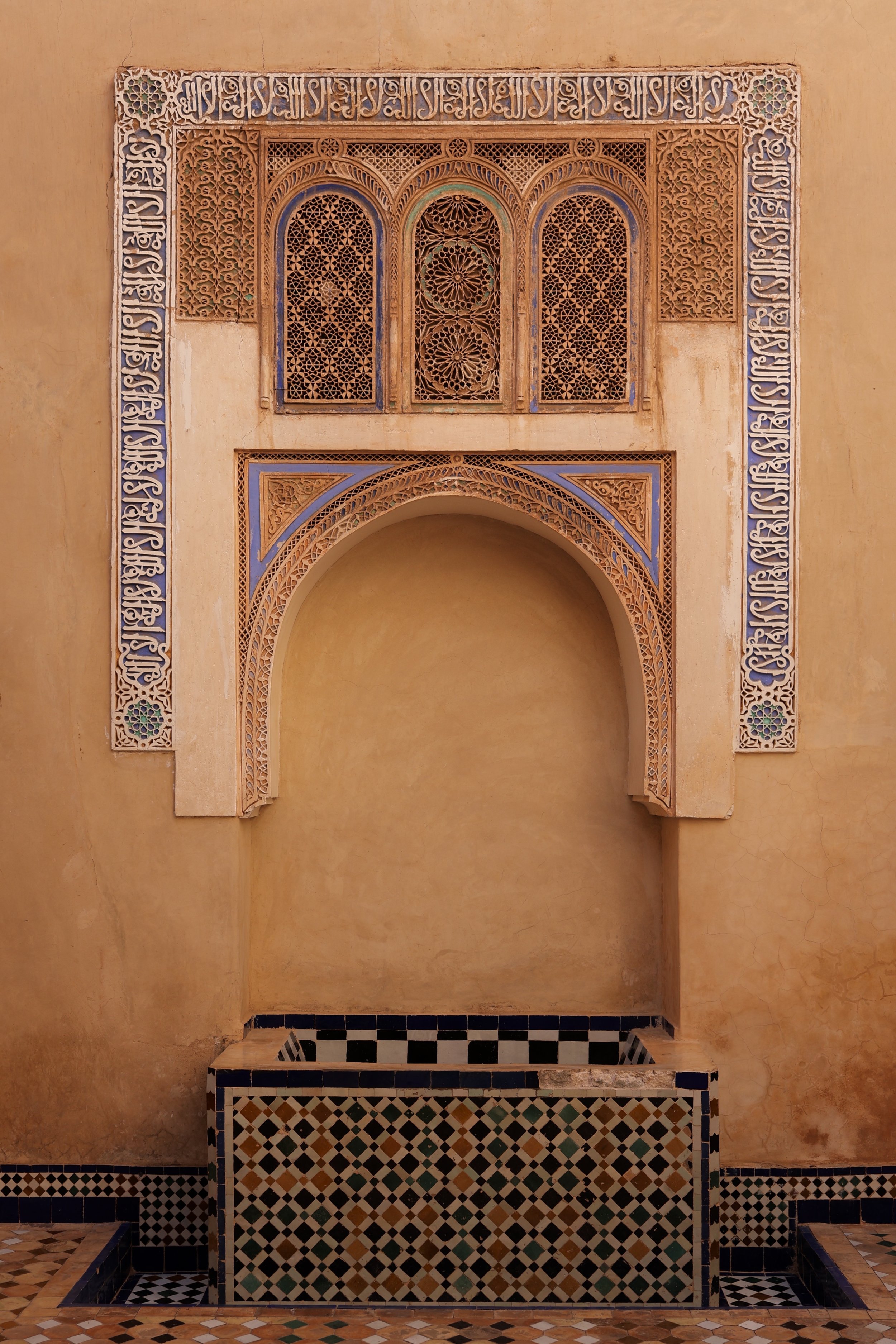 Decorative fountain with intricate Arabic geometric design on the wall above, set in a warm-toned wall, featuring a tiled basin in green, black, and beige. In Marokko.