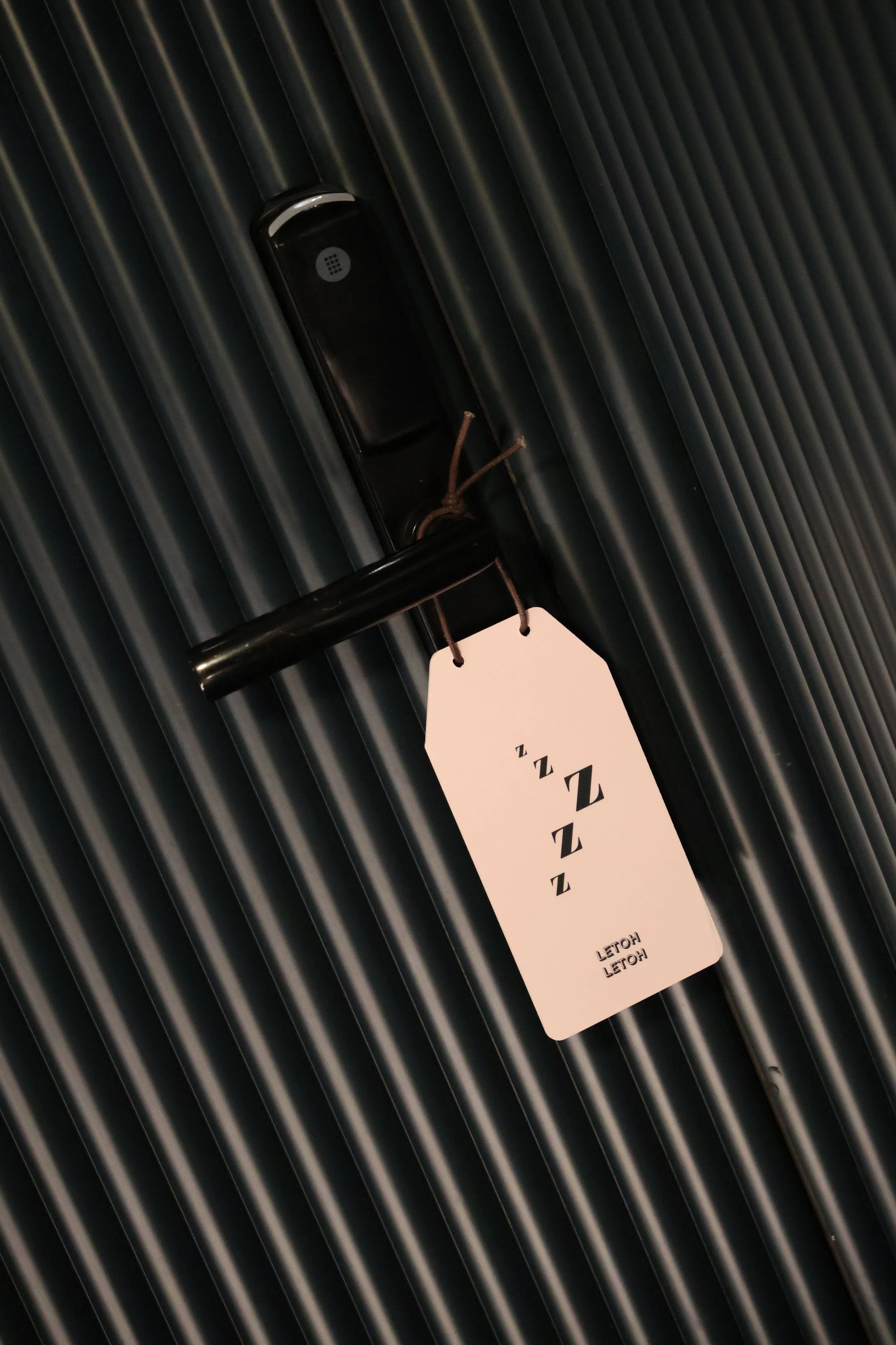 A black door handle with a white tag hanging from it, displaying a series of 'Z' symbols and the text 'LETOH LETOH HOTEL' on a dark, vertically ridged door.