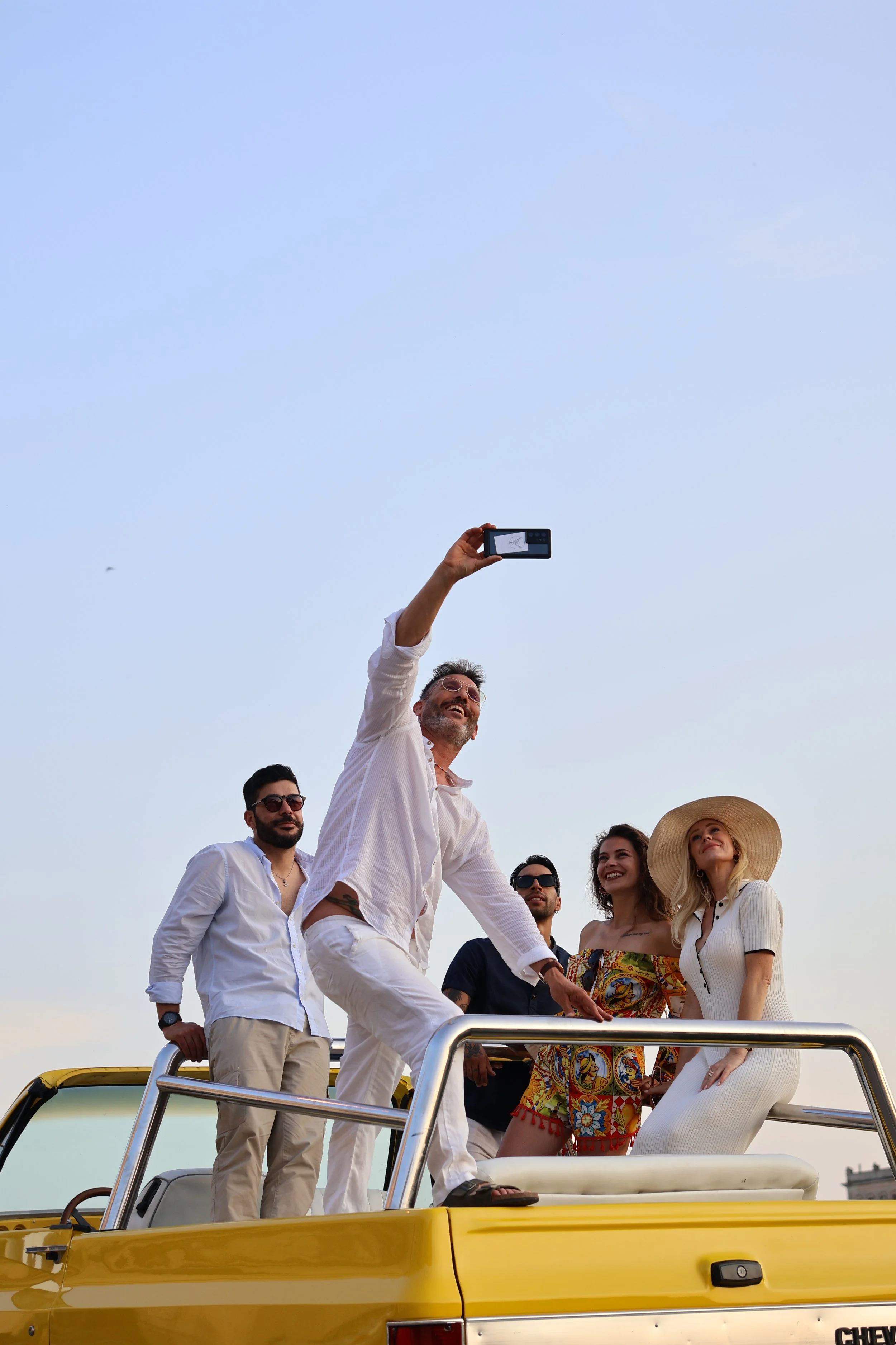 A group of five friends taking a selfie on top of a yellow convertible car during sunset, dressed in summer clothes, with a clear blue sky in the background.