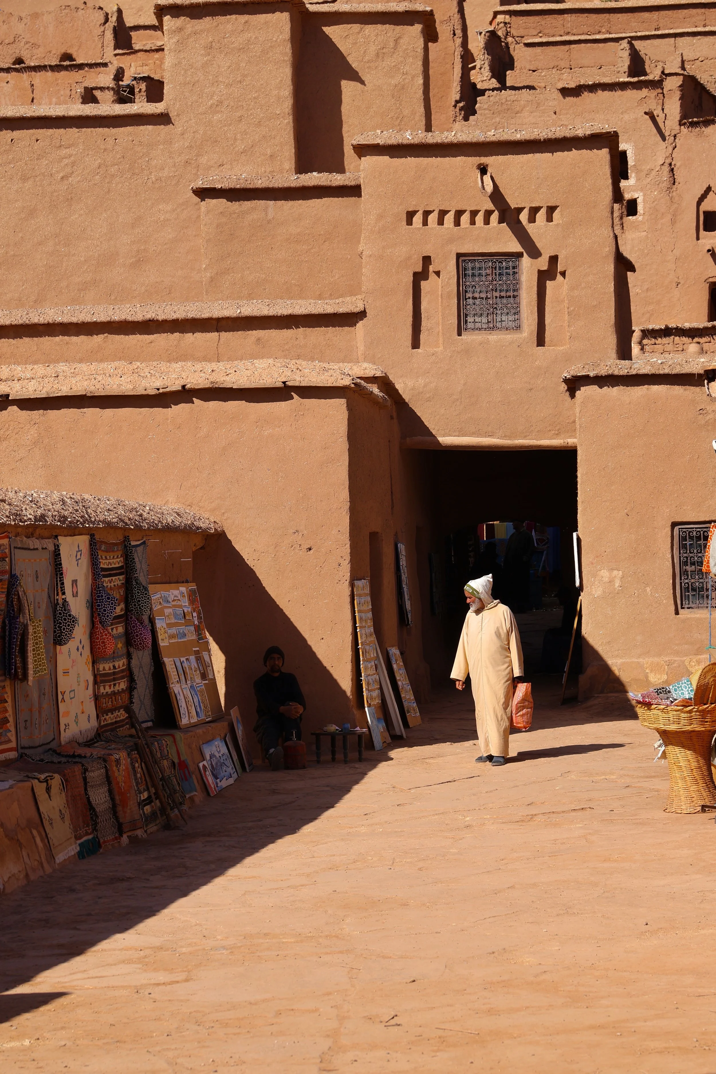 People walking and selling handmade textiles and artwork under a large adobe archway in a desert town. In Marokko.