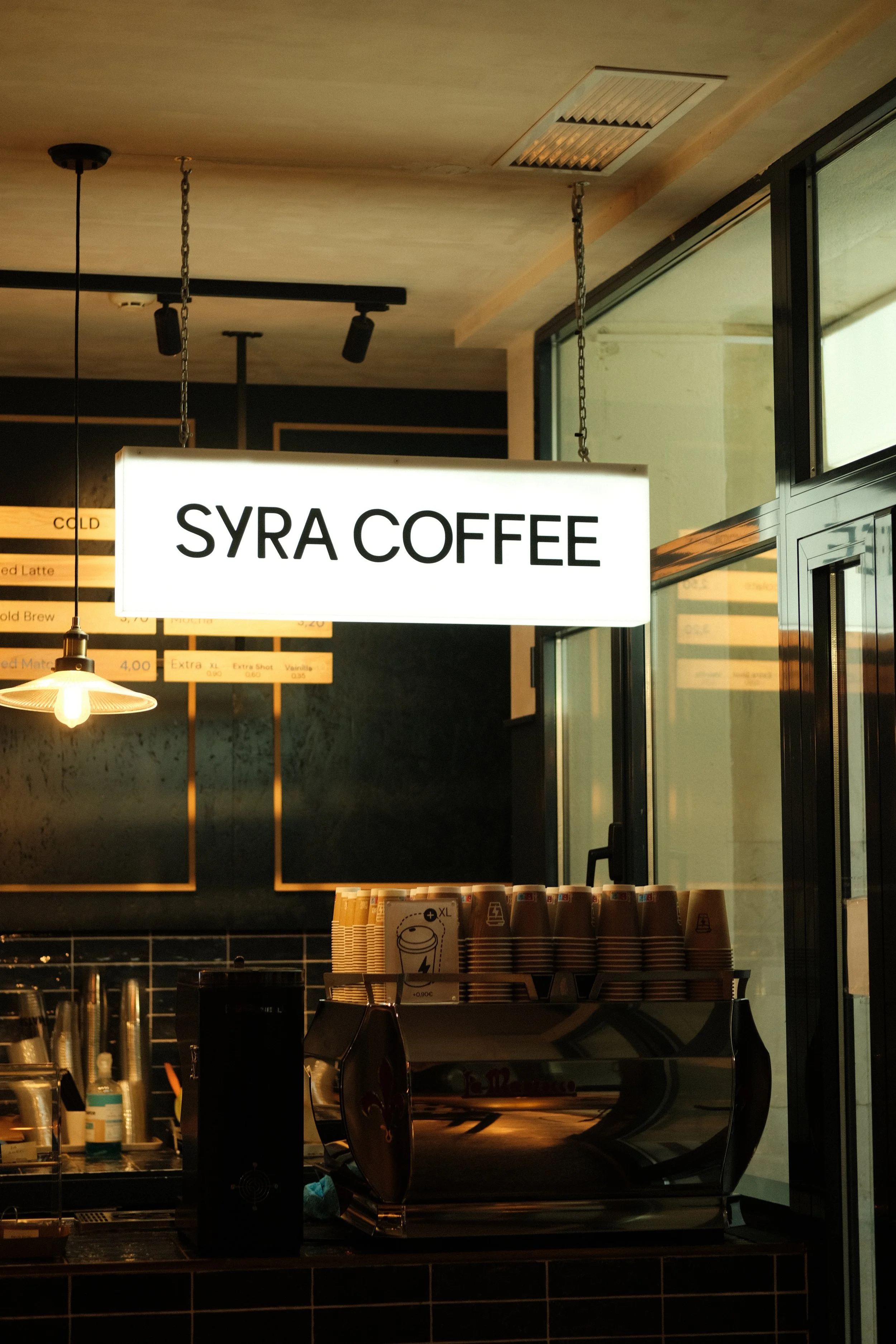 A brightly lit coffee shop with a sign that reads 'SYRA COFFEE' hanging from the ceiling near the large windows.