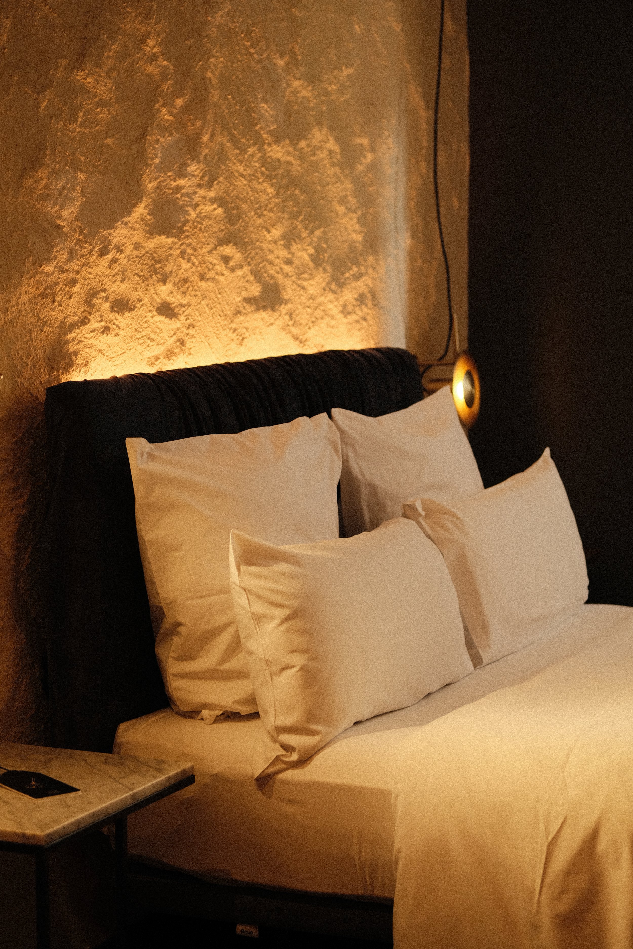 A cozy bedroom with a bed featuring white pillows and linens, a textured wall illuminated by warm lighting, a round wall-mounted lamp, and a small marble-top side table.