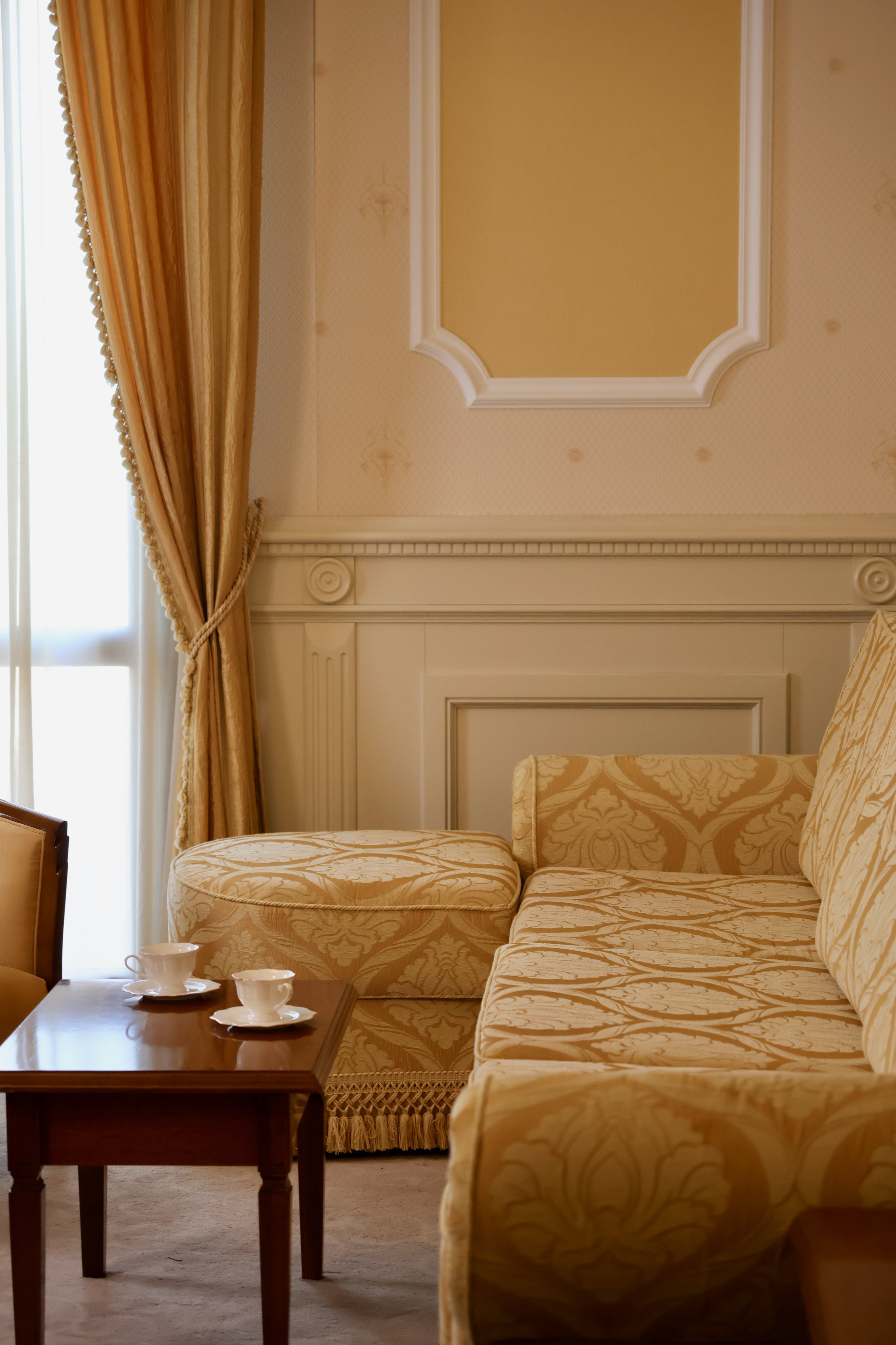 Elegant hotel room with yellow patterned sofa, matching ottoman, wooden side table with two cups and saucers, cream-colored walls with decorative molding, and gold curtains by a large window.