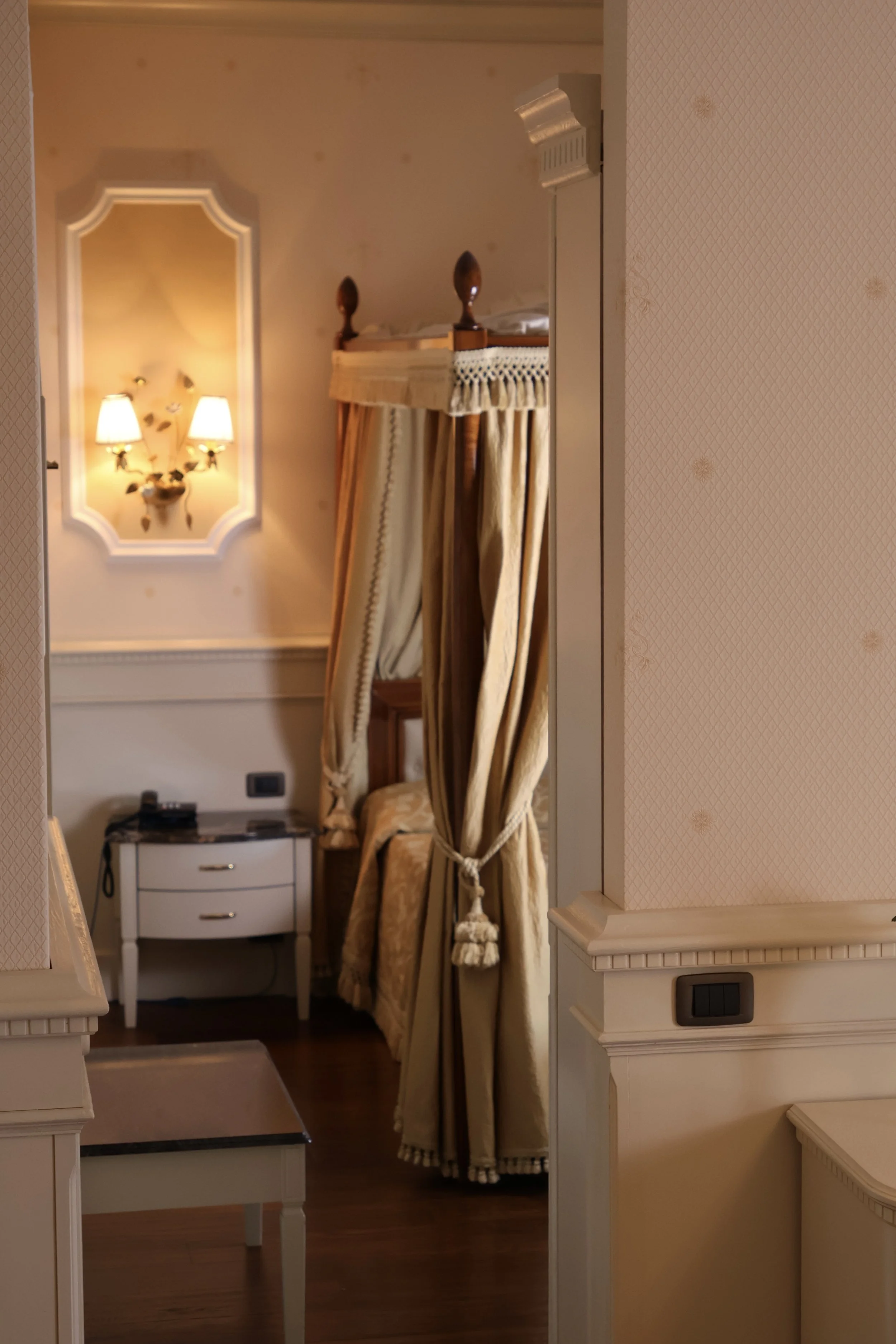 A view of a hotel room corner with a four-poster bed with beige curtains, a small white bedside table, and decorative wall lamps with yellow shades.
