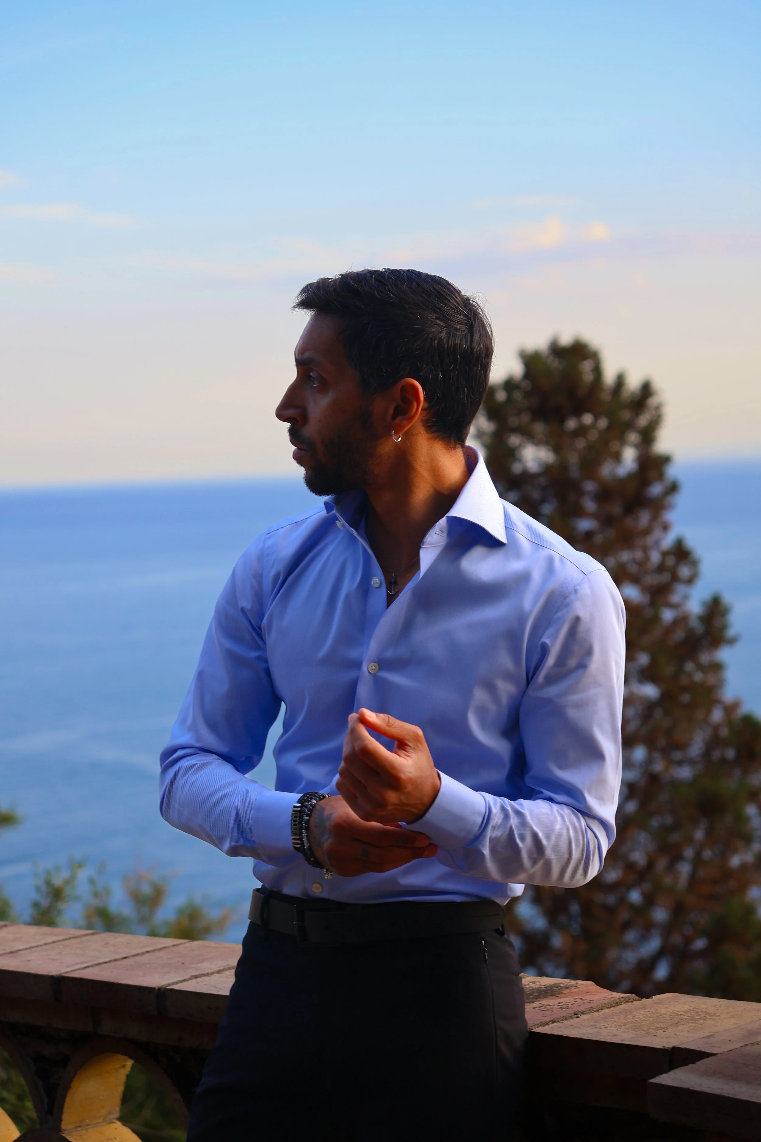Man in a white shirt standing outdoors with ocean, trees, and sky in the background.