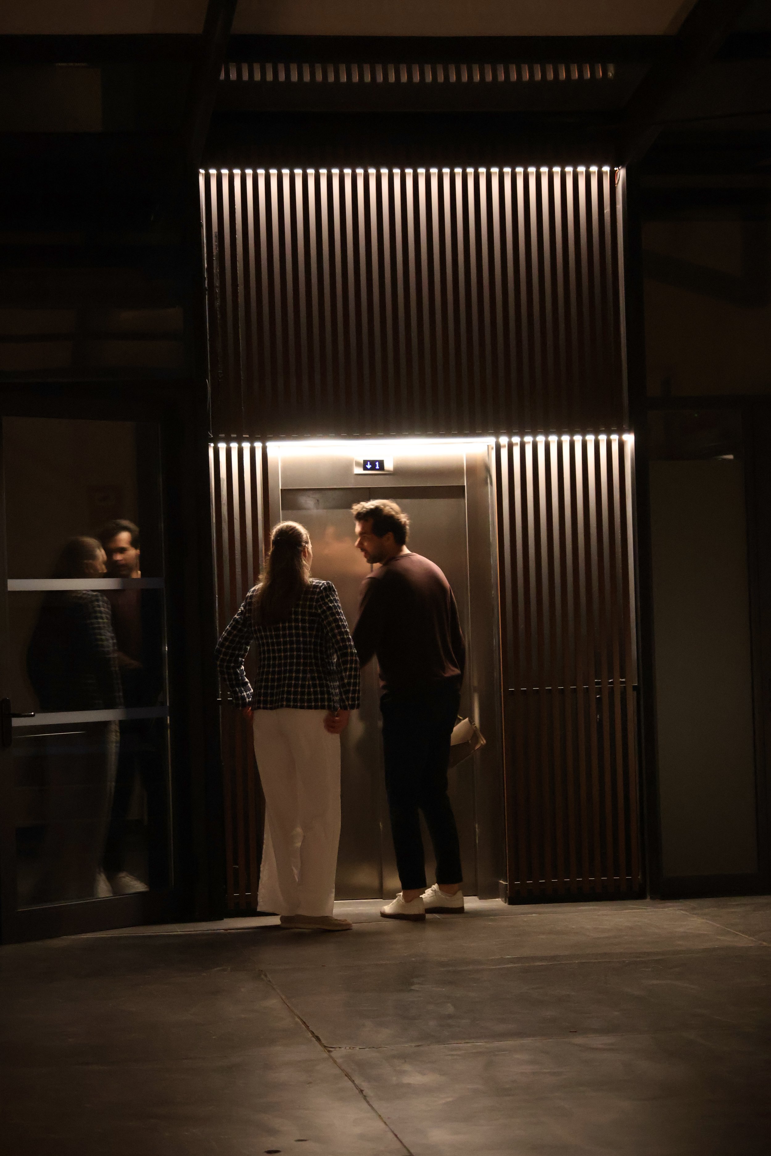 Two people talking near an elevator in a dimly lit building.