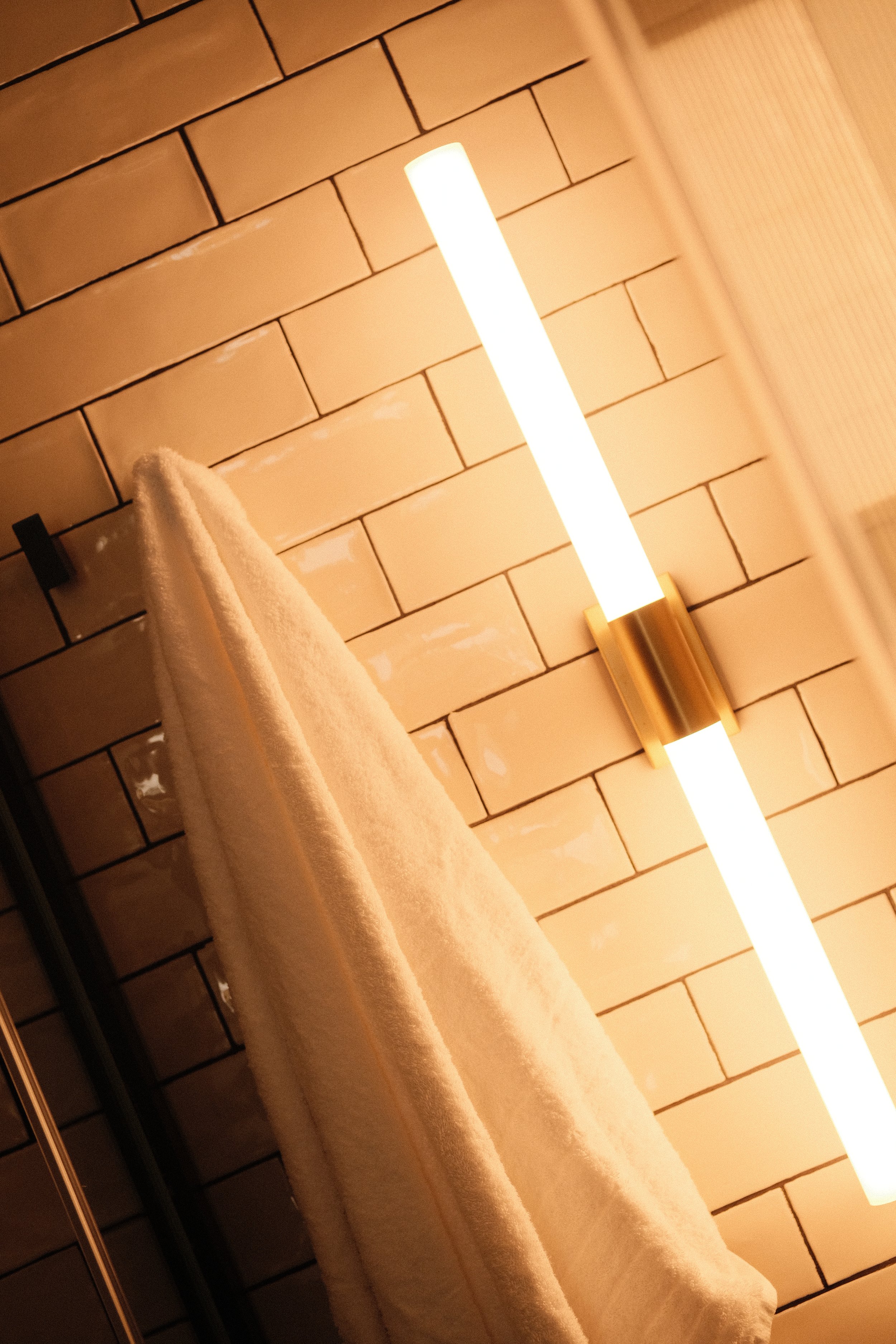 A white towel hanging on a black hook on a beige brick wall with a modern vertical light fixture.