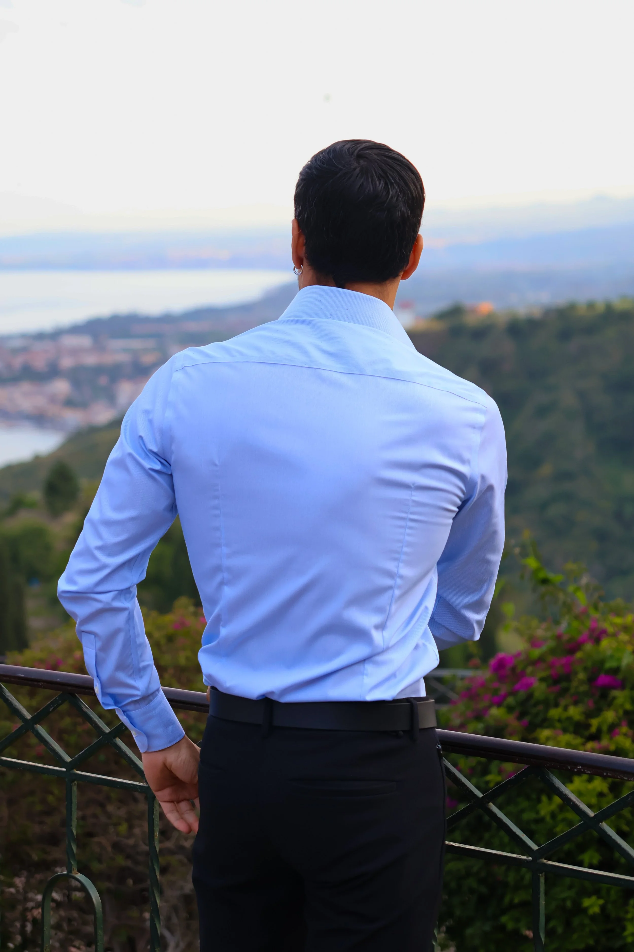 A man wearing a light blue dress shirt and black pants standing by a railing, looking out over a landscape with greenery, hills, and water in the distance.