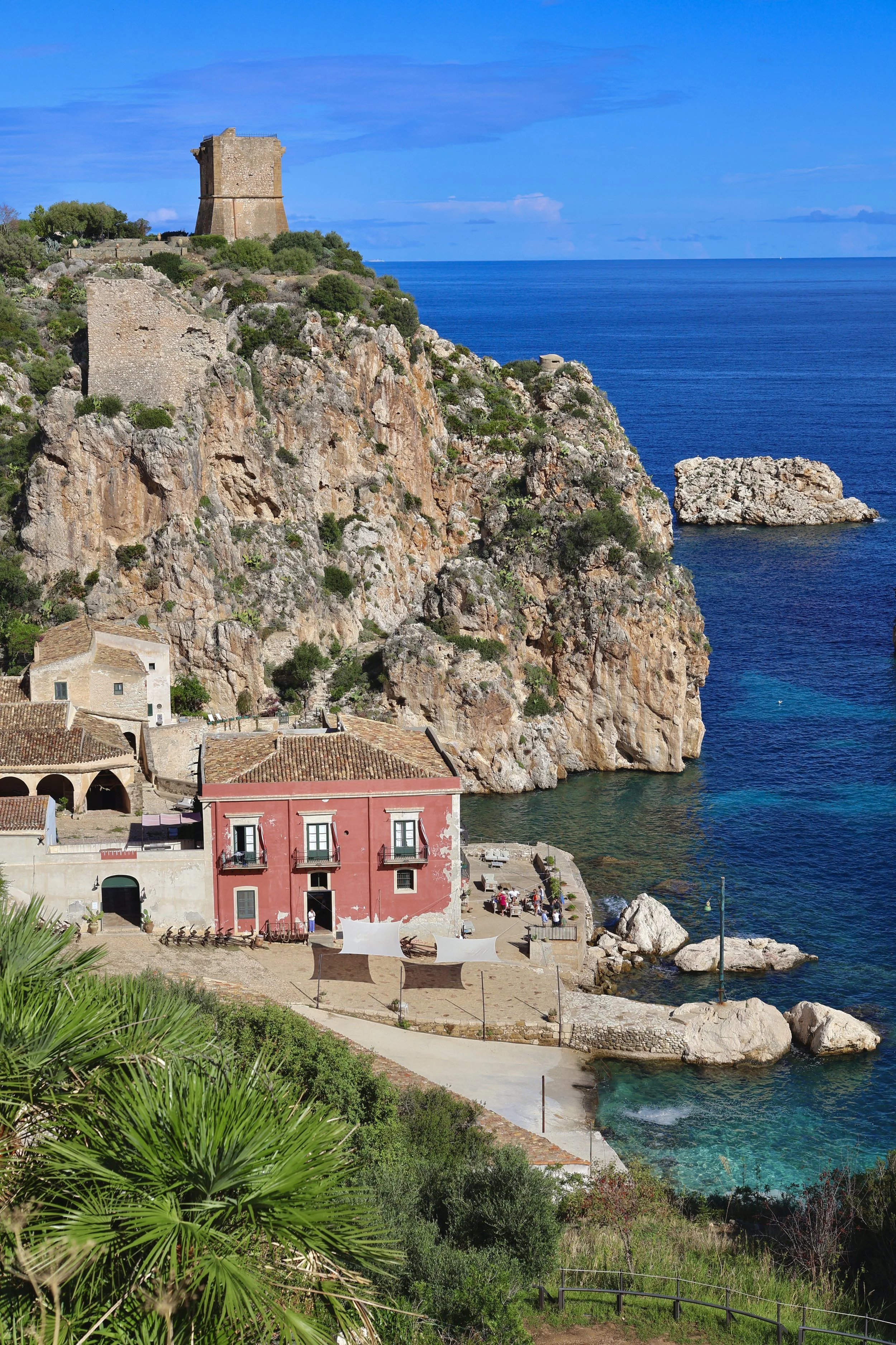 Scenic view of a coastal village with colorful buildings, rocky cliffs, and a deep blue sea under a partly cloudy sky. in Scopello, Sicily