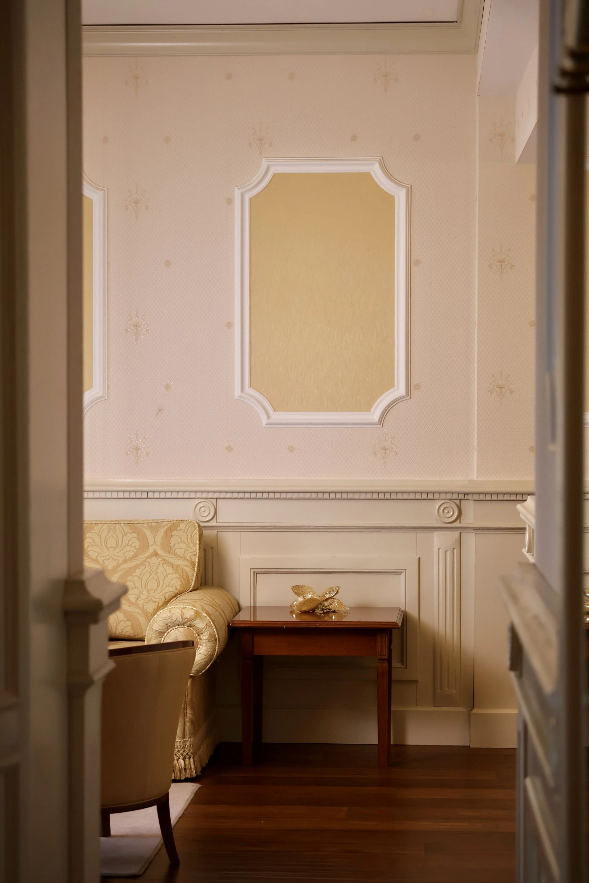 View of an elegant, vintage-style room with a beige sofa, a small wooden table with a decorative gold-colored object, and cream-colored paneled walls with decorative molding.