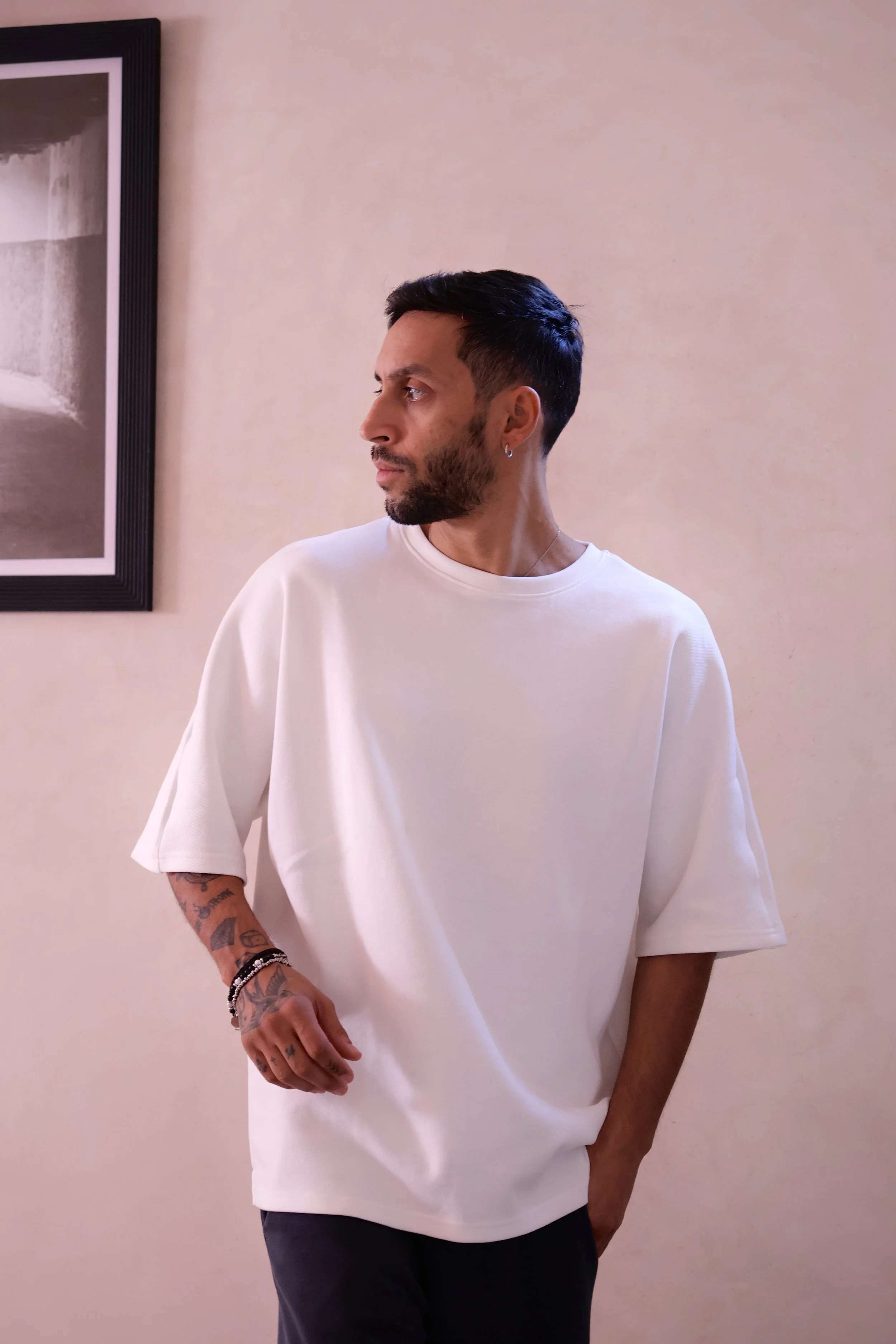 Editorial foto for men's fashion. A man with short dark hair and tattoos on his arm, wearing a white oversized t-shirt, standing indoors against a plain beige wall, looking to his left.