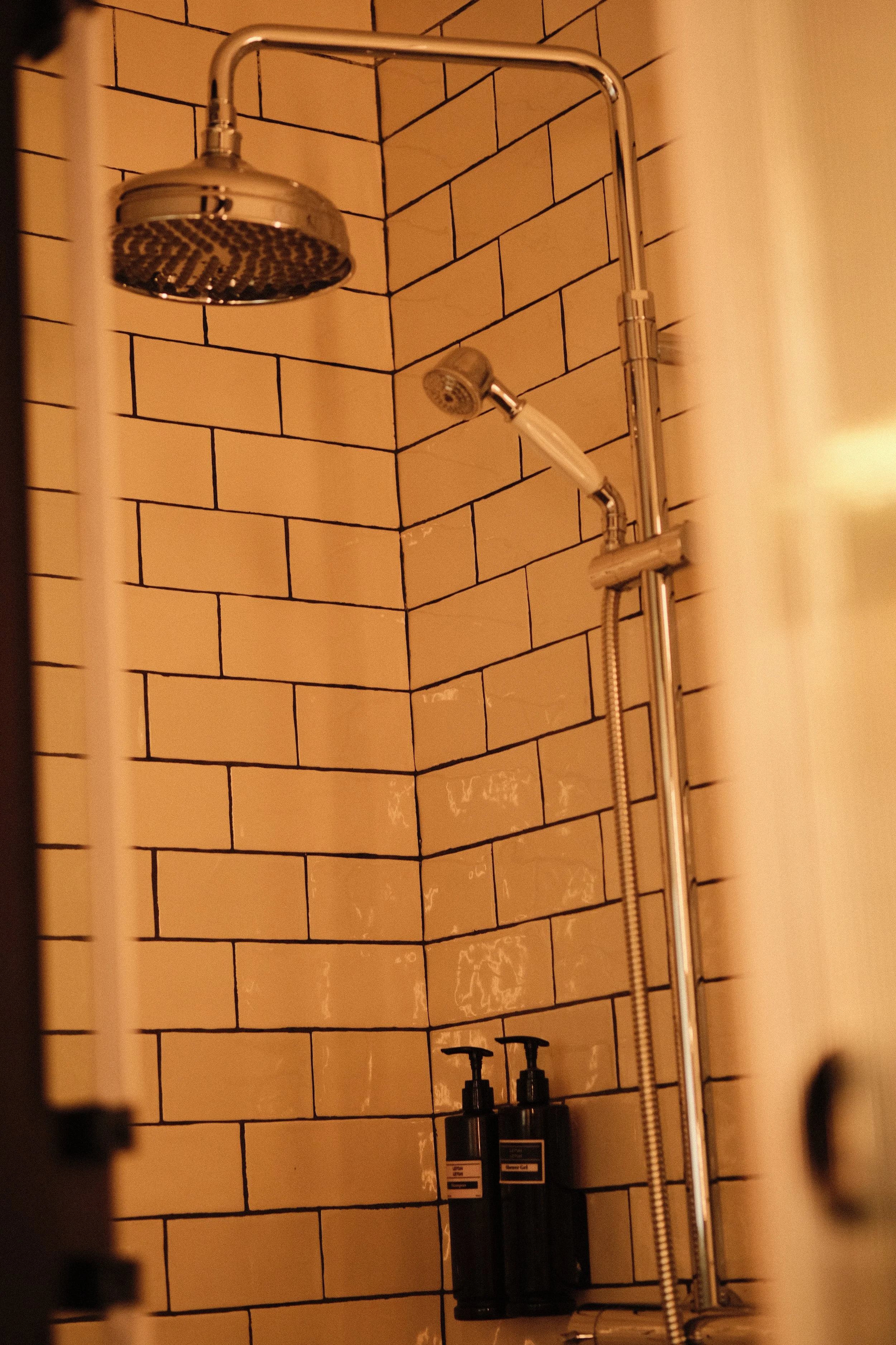 Shower with beige subway tiles, metal showerhead, handheld shower, and two black bottles on a corner shelf.