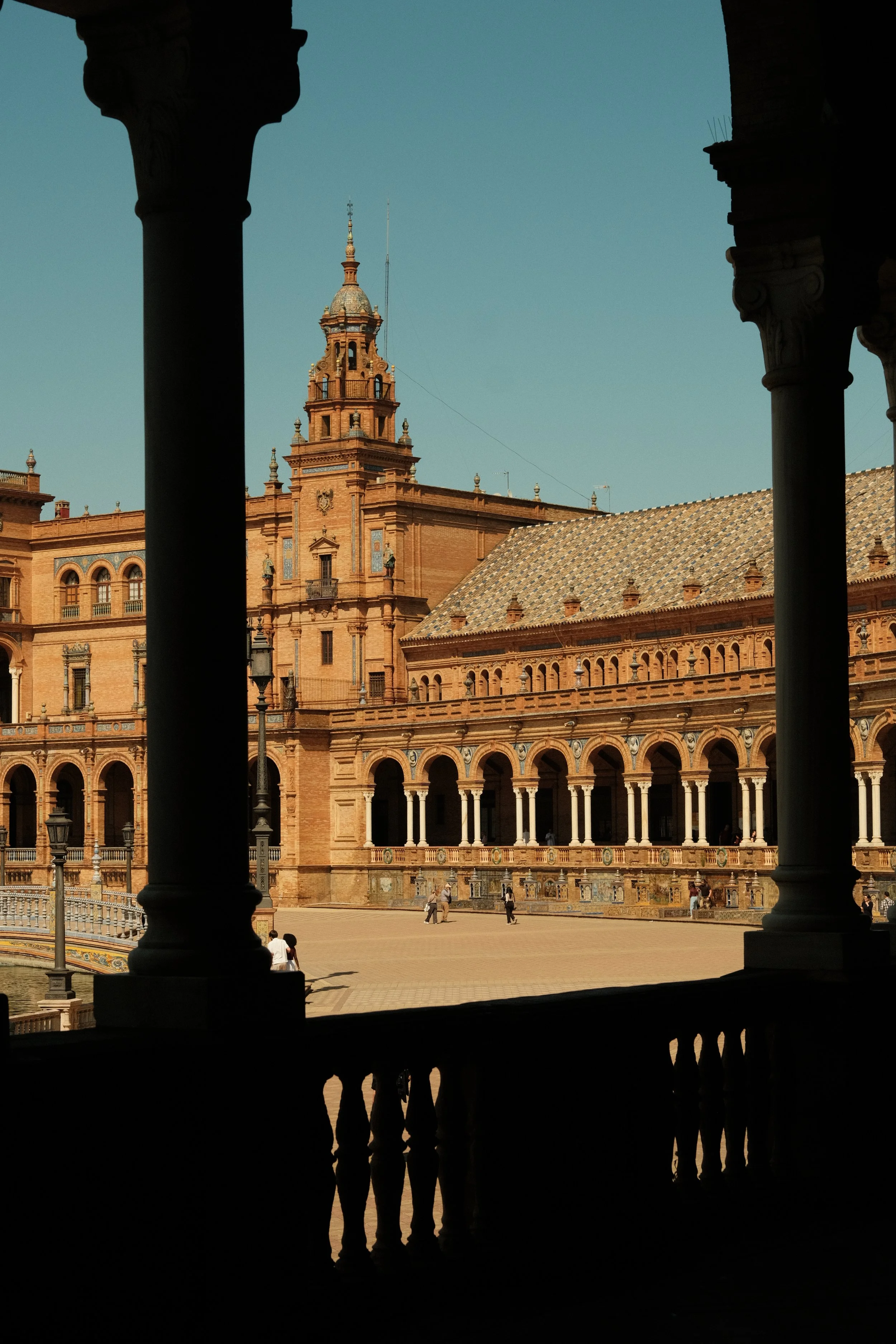 View of a historical building with a tower, seen through a dark inner structure, with a clear sky in the background and a few people walking in the open courtyard.
